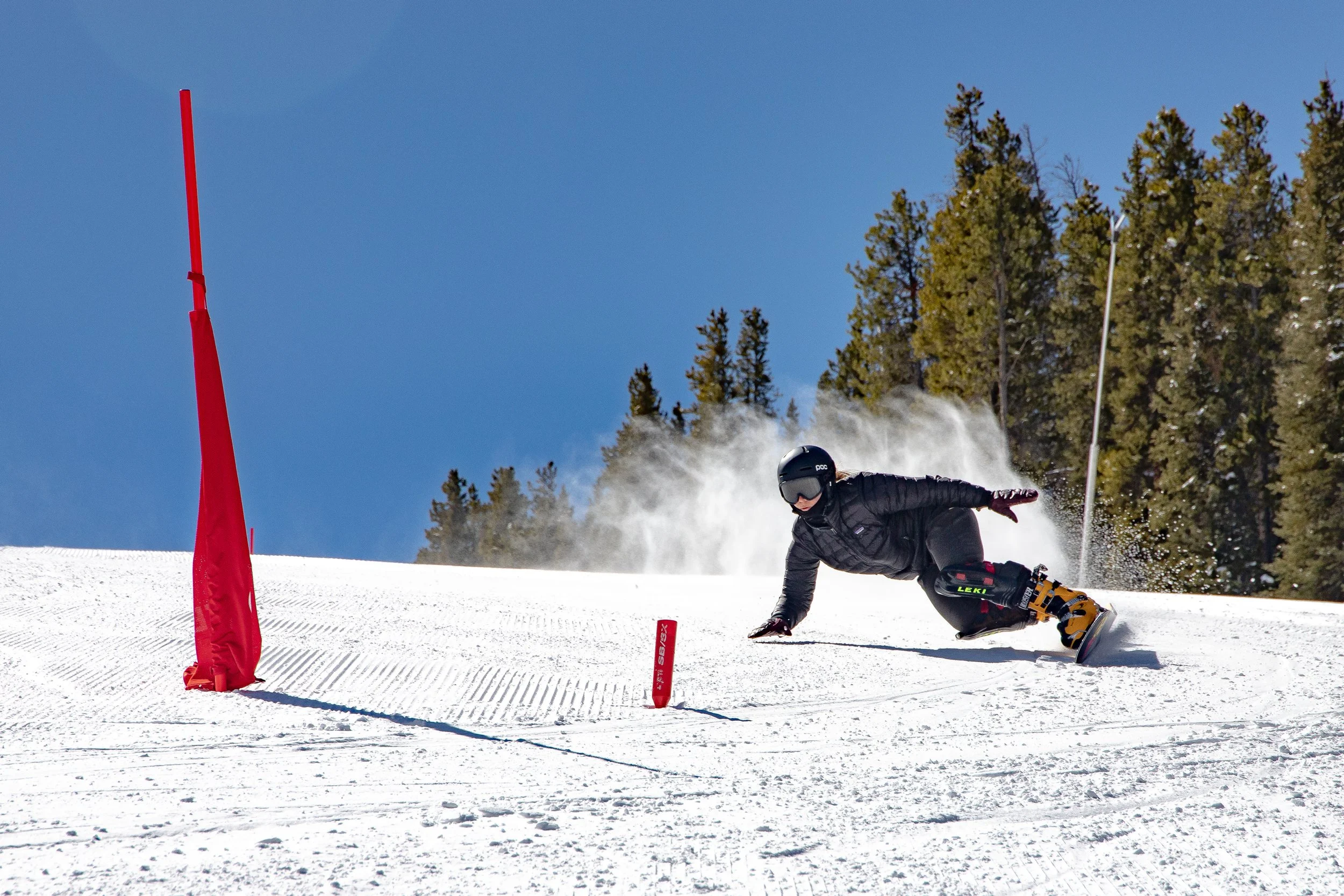 A person in black winter gear and a helmet snowboarding down a snow-covered slope around red gate flags on a clear, sunny day with trees in the background.