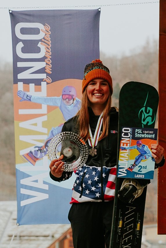 A woman in winter clothes, holding a snowboard, a medal around her neck, and a trophy, standing in front of a banner for the Valemoto Junior World Championships.