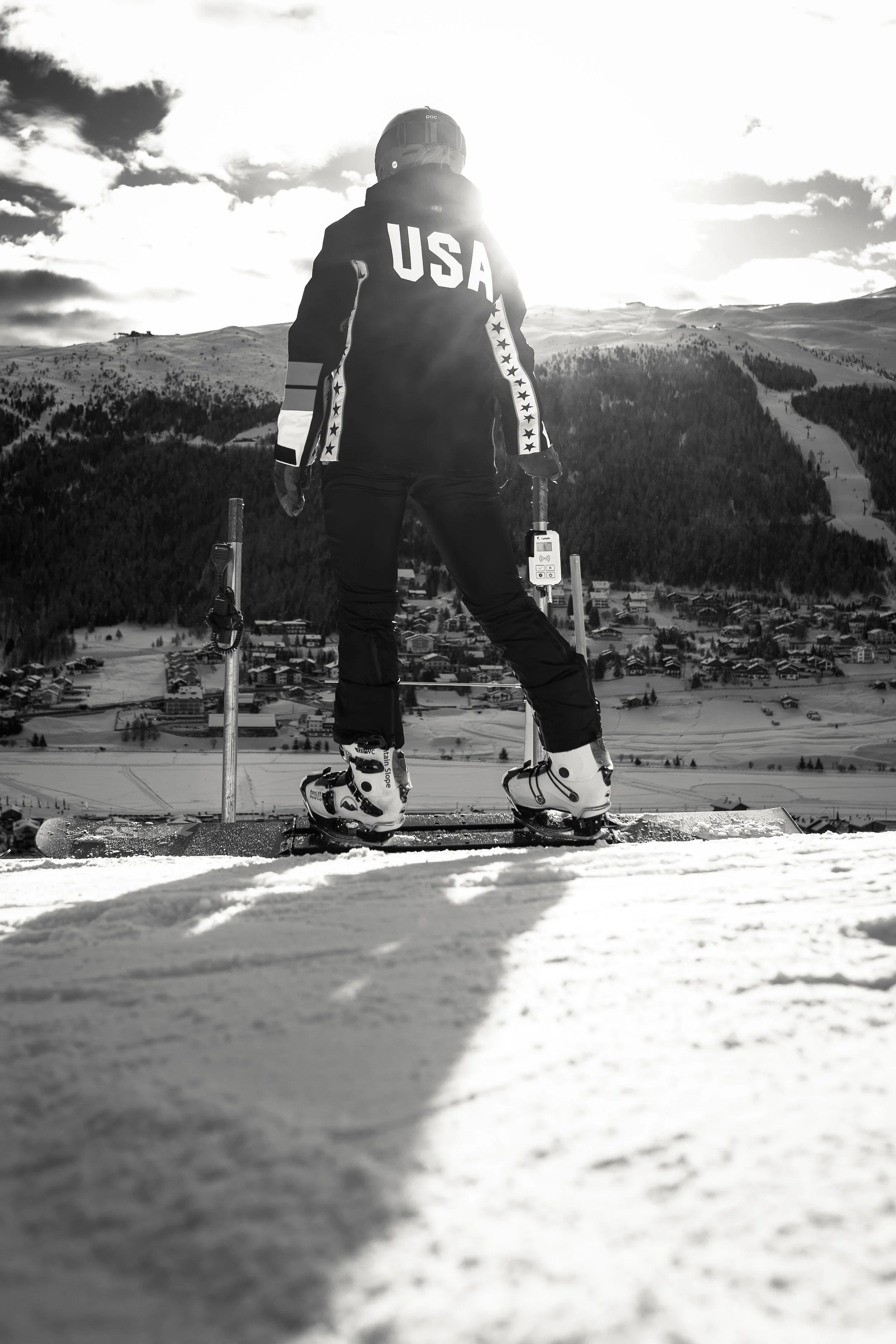 A skier standing on snow with ski poles in hand, facing away towards a mountainous landscape, wearing a jacket with 'USA' on the back, during daytime with partly cloudy sky.