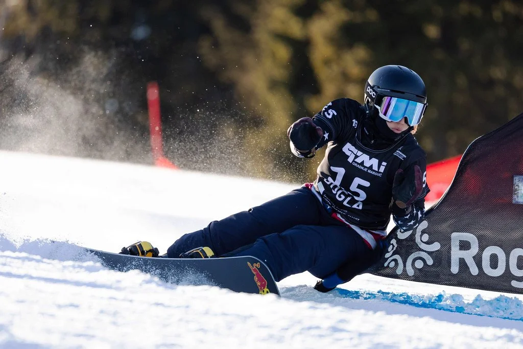 A person racing on a snowboard, wearing a black helmet and goggles, leaning into a turn on a snowy slope during a competition.
