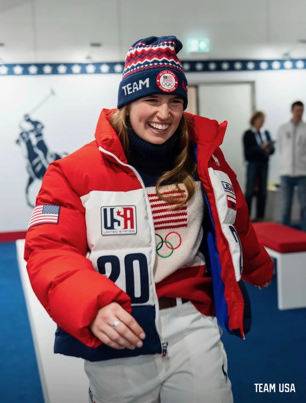 Smiling female athlete in Team USA winter sports outfit, wearing a USA beanie, red jacket, and Olympic gear, standing in an indoor sports facility.