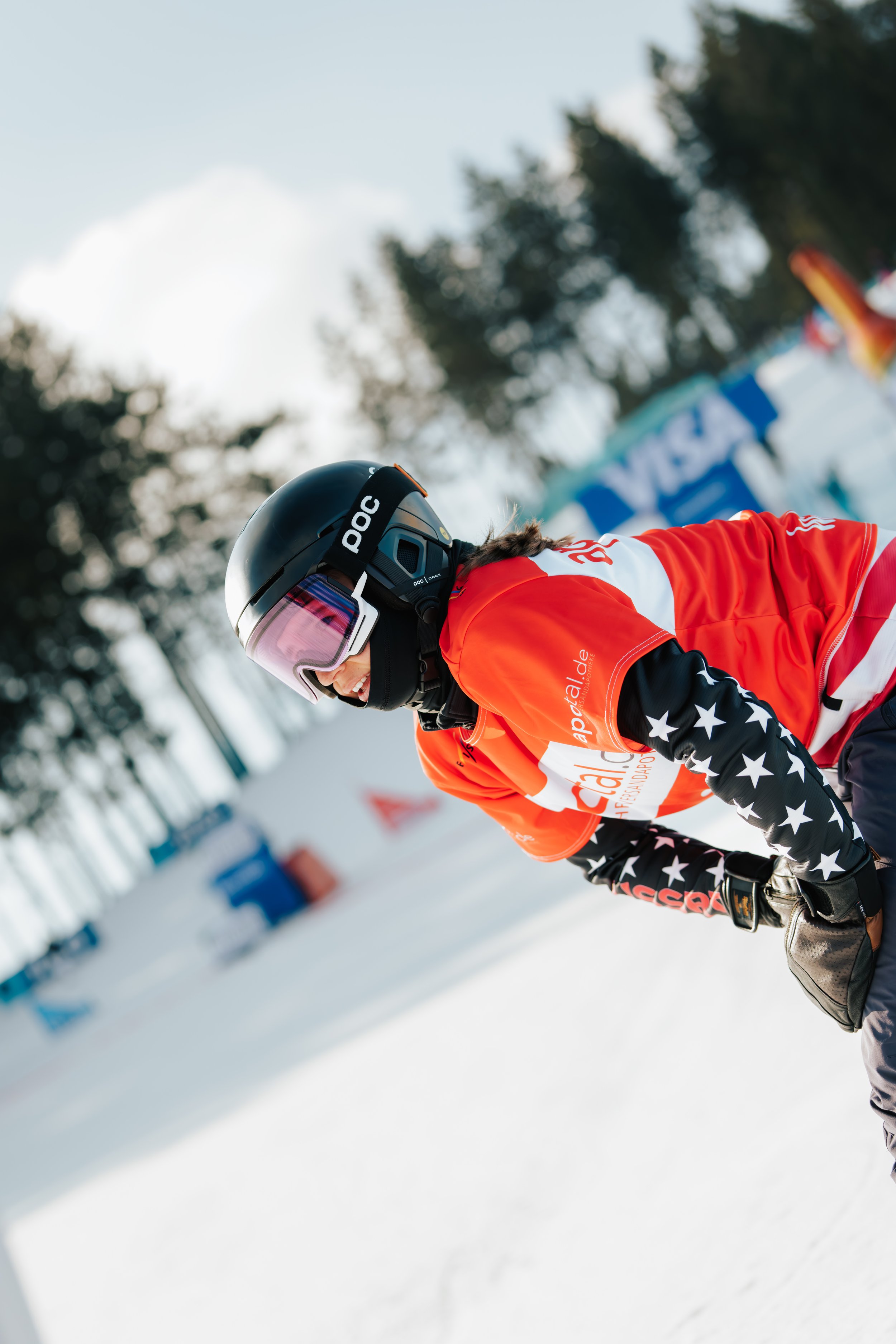 A person in a black helmet and bright orange ski jacket with star patterns on the sleeves, leaning forward on skis in a snowy outdoor setting, with blurred trees and banners in the background.