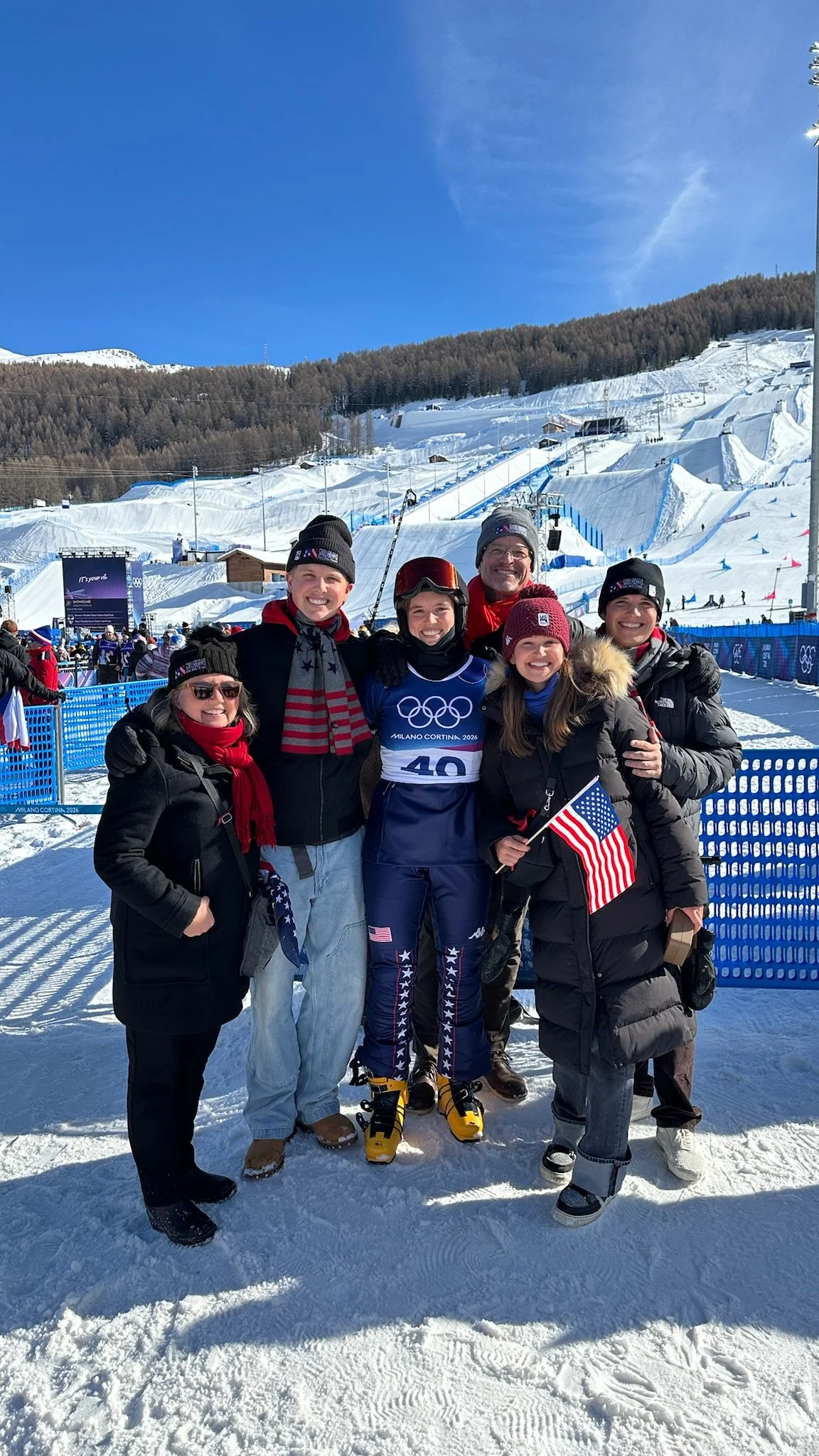 Group of six people standing in snowy ski resort with ski slopes and trees in the background, smiling and celebrating, some holding American flags, wearing winter jackets, hats, and ski gear, during daytime.
