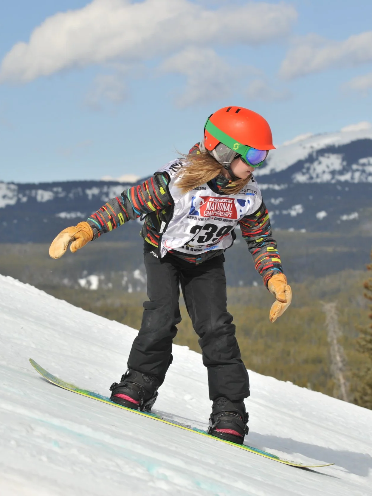 Young girl skiing downhill on snow with mountains in background, wearing orange helmet, colorful jacket, goggles, and gloves.