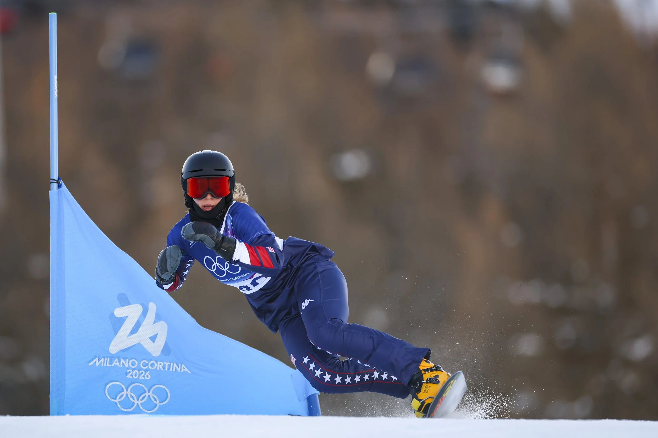A snowboarder wearing a black helmet, red goggles, a blue jacket, and yellow boots, carving down a slope at the Milano Cortina 2026 Winter Olympics. The snowboarder is leaning into a turn near a blue gate with the Olympic rings. 