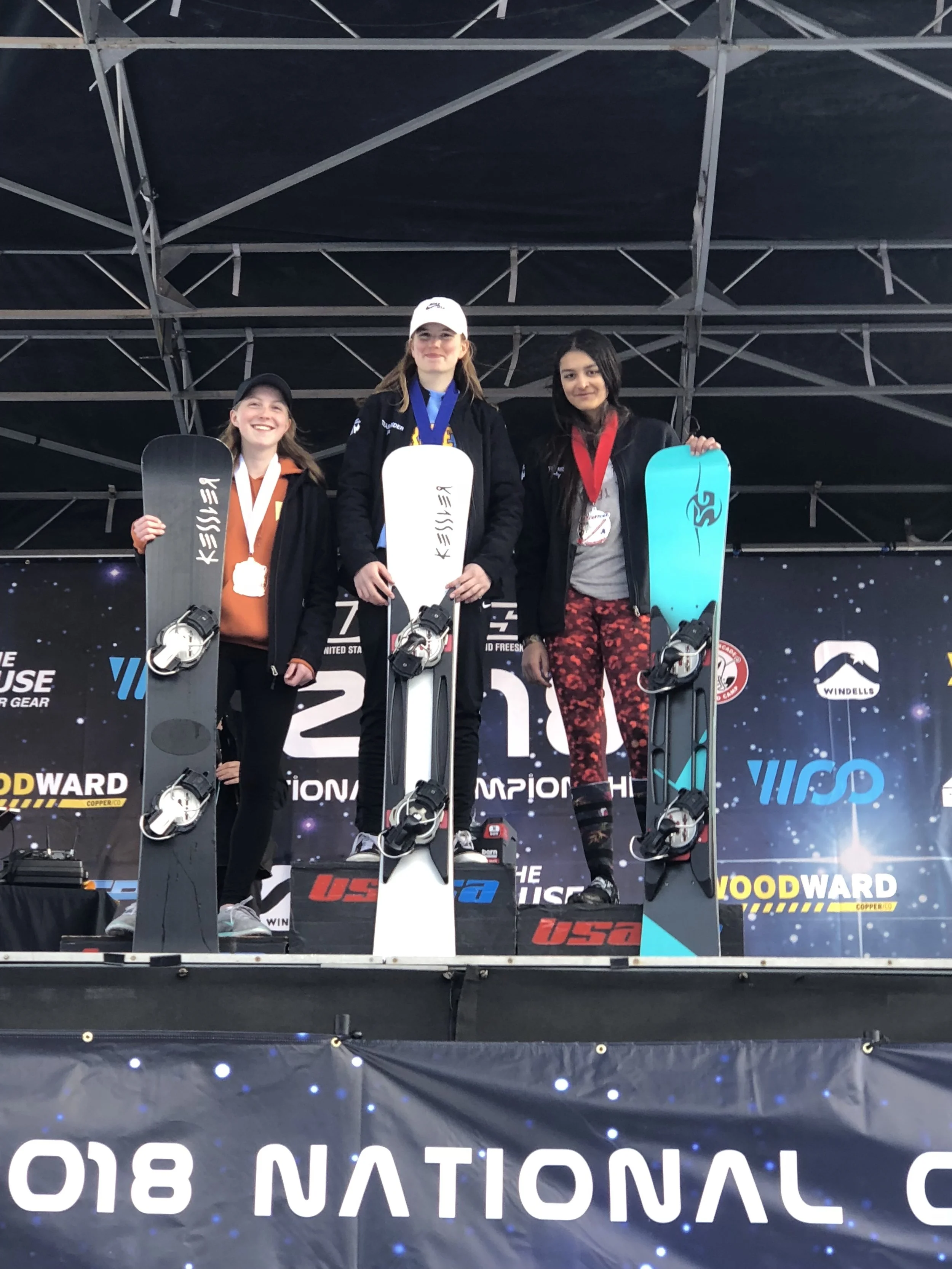 Three women standing on a winners' podium at a snowboarding competition, each holding a snowboard and wearing medals. The woman in the center is on the first-place podium, wearing a white cap and black jacket, with a gold medal. The woman on the left is on the second-place podium, wearing a black hat and orange shirt, with a silver medal. The woman on the right is on the third-place podium, wearing a gray shirt and black jacket, with a bronze medal.