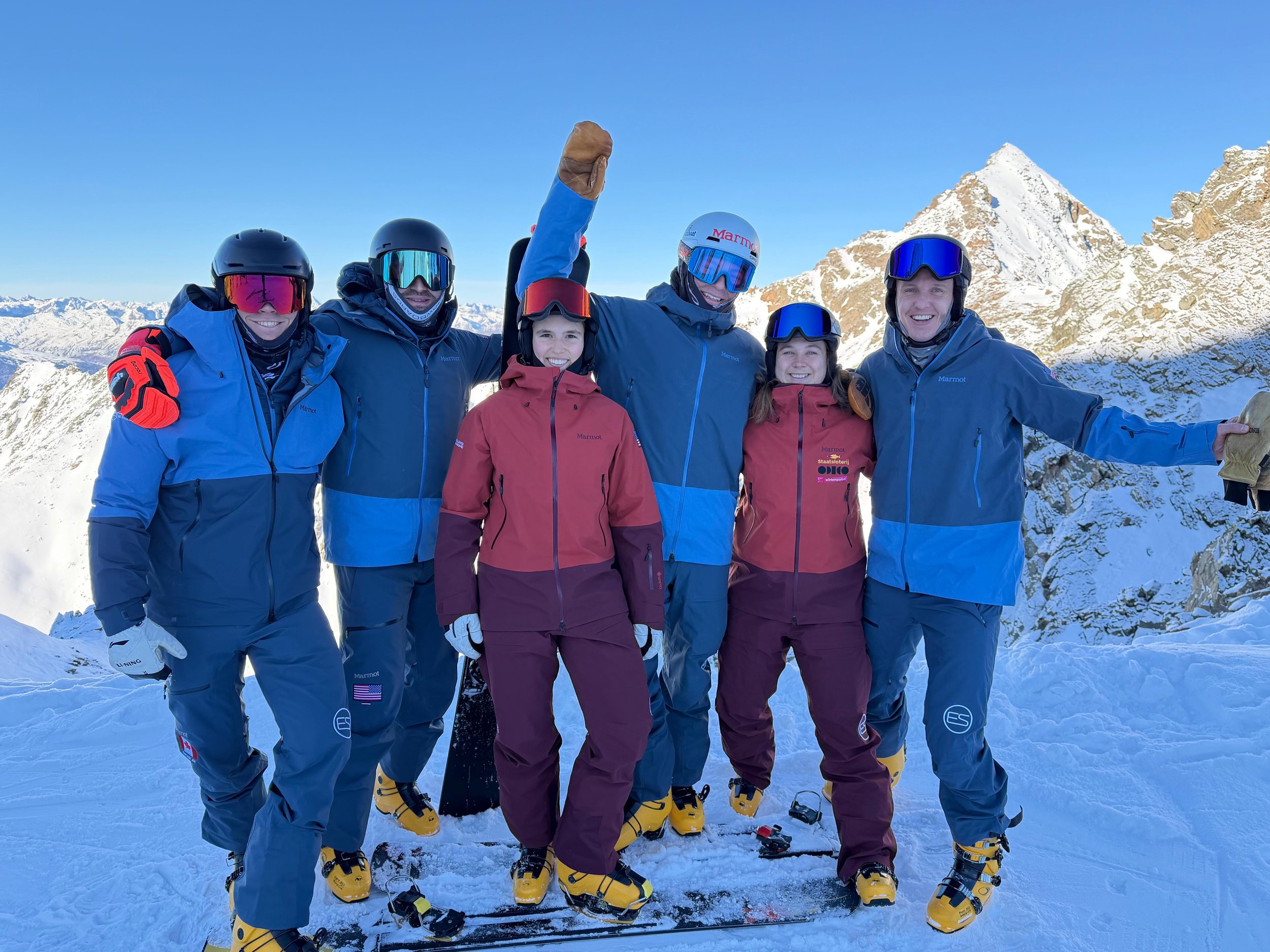 Group of six snowboarders posing on a snowy mountain with snow-covered peaks in the background. They are wearing blue and red Marmot jackets. Some have their arms around each other, smiling and celebrating.