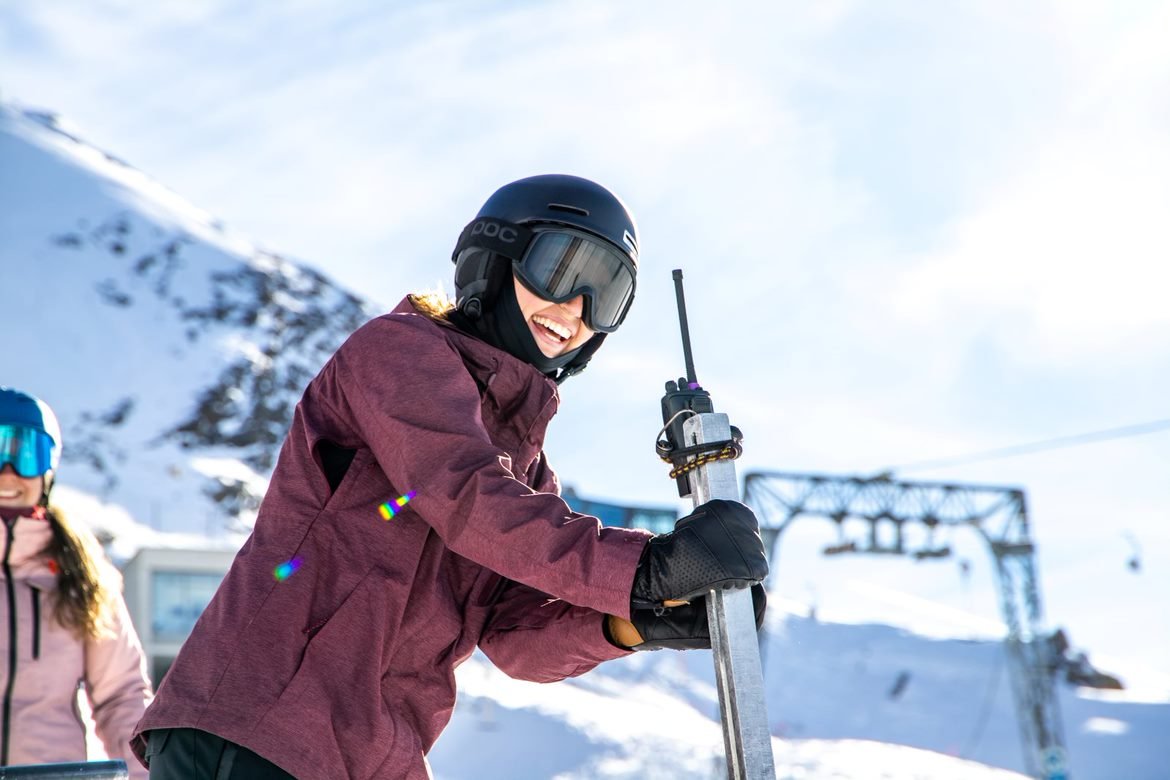 Person in a maroon ski jacket with a black helmet and goggles smiling and speaking into a radio at the start gate on the mountain.
