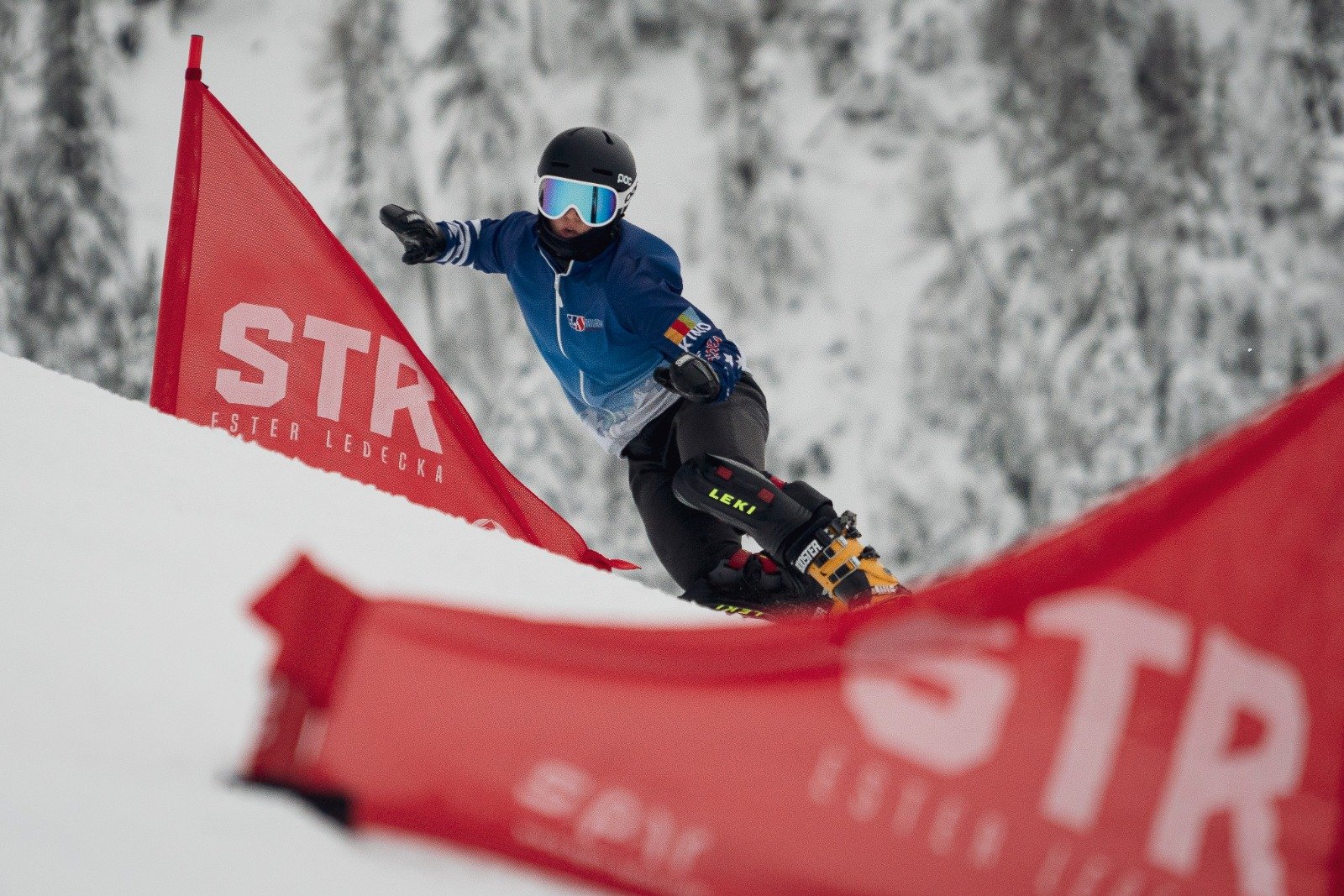A snowboarder in a blue jacket and black helmet riding around red pannels on a snowy slope with snow-covered trees in the background.