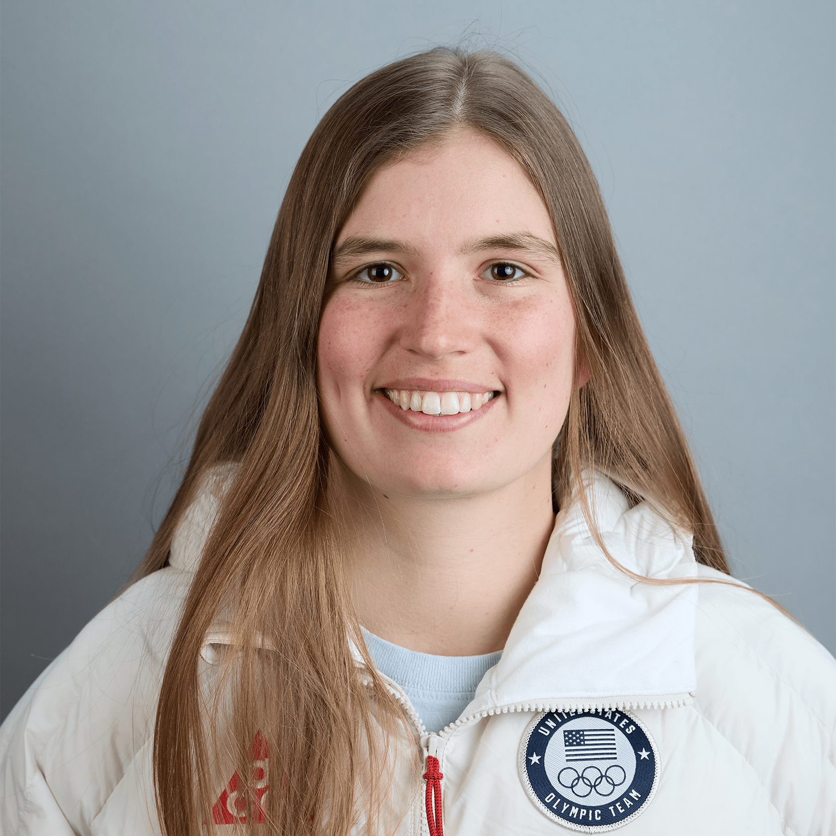 A young woman with long light brown hair smiling, wearing a white jacket with an Olympics team patch on the chest, against a gray background.