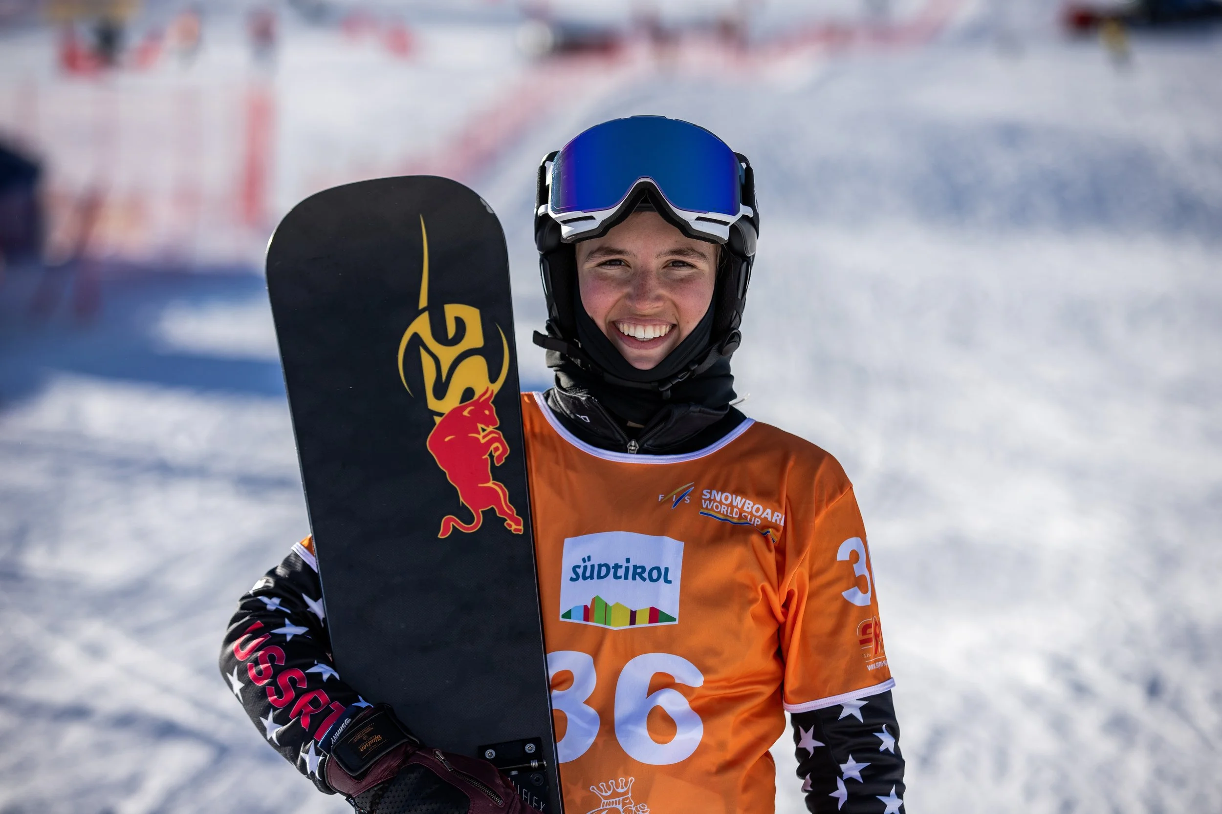 A young female snowboarder smiling while holding a SG snowboard on a snowy mountain, wearing an orange jersey with the number 36. A post race photo. 