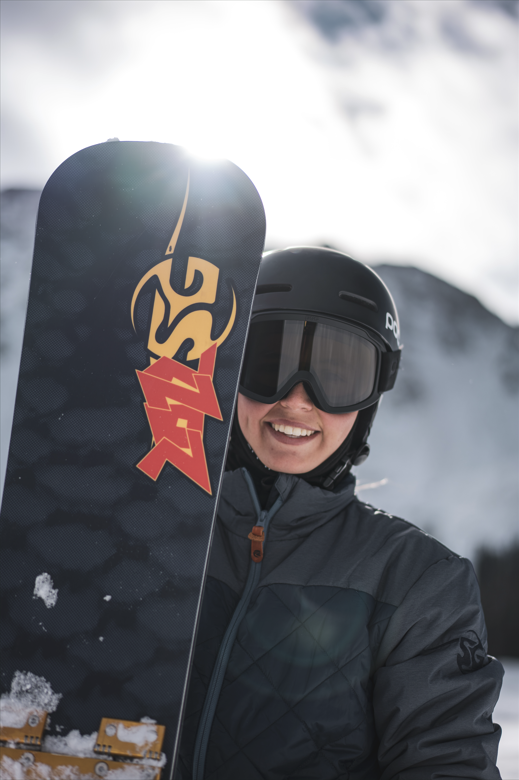 Smiling person in snowboard gear holding a SG snowboard with a mountain background.