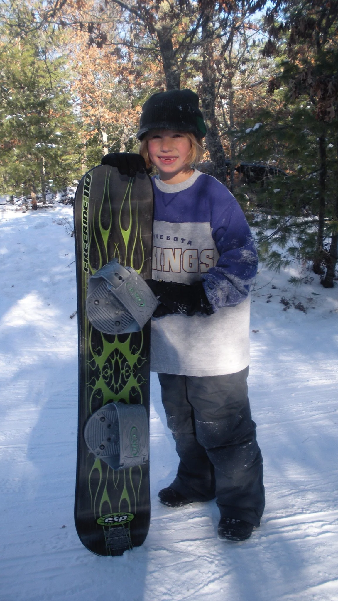 A child dressed in winter clothes standing on snow, holding a snowboard, outdoors in a wooded area with trees and sunlight.