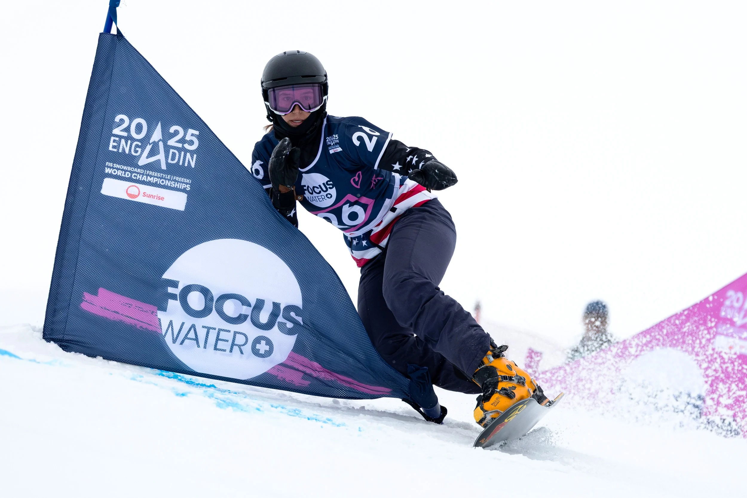 A snowboarder carving on snow next to a large blue gate with white and pink text that reads 'FIS Snowboard | Freestyle | Freeski World Championships'. 