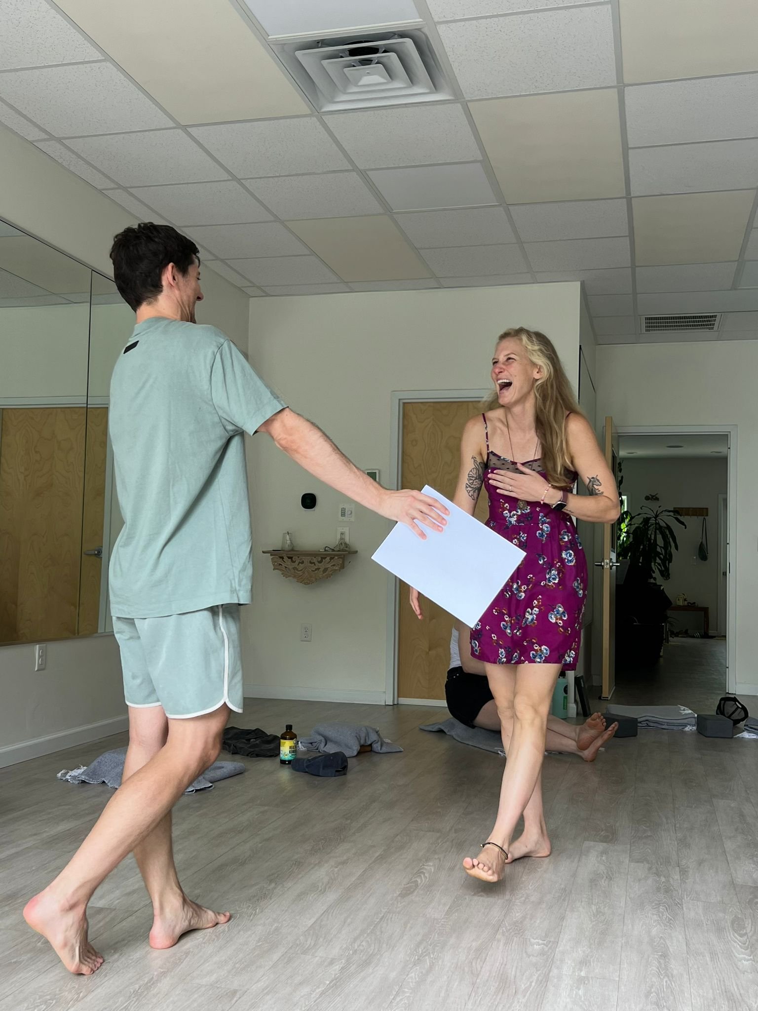 A woman in a floral dress and tattoos on her arm is smiling and laughing while dancing. She is barefoot and holding a piece of paper. A man in casual clothing is standing nearby, reaching out to her. There is another person lying on the floor in the background, surrounded by various items.
