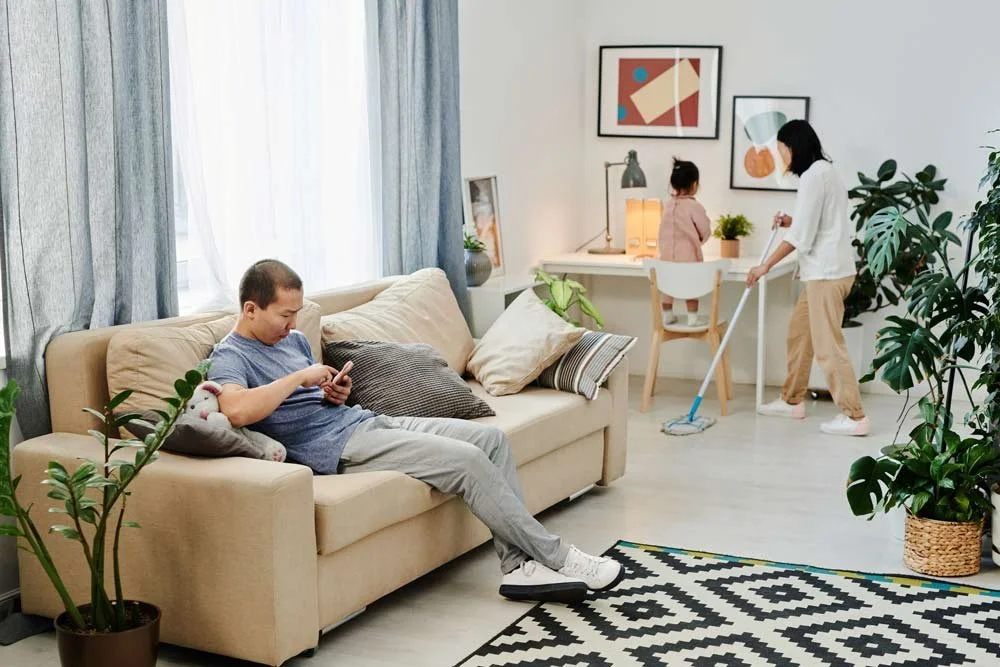 Living room scene with a man sitting on a beige sofa using his phone, a woman cleaning the floor with a mop, and a young girl sitting at a desk with plants and framed artwork.