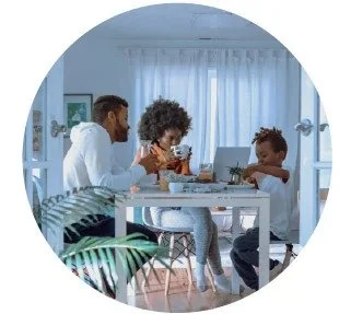 A family of three sitting at a dining table in a bright, airy room with white curtains, enjoying breakfast together.