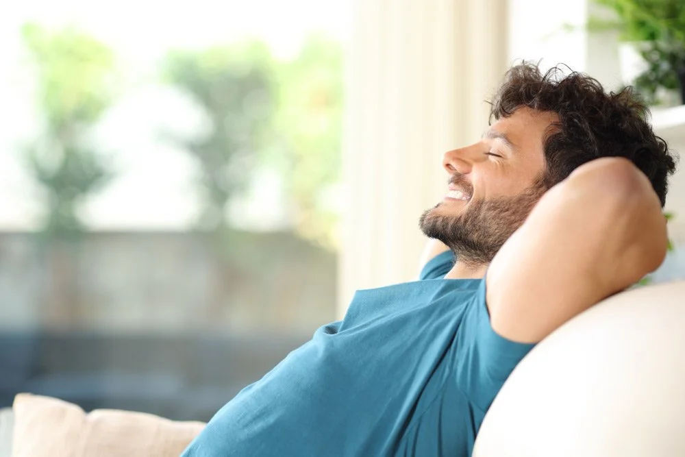 A man relaxing on a couch with eyes closed, smiling, wearing a blue t-shirt in a bright room with large windows and greenery outside.