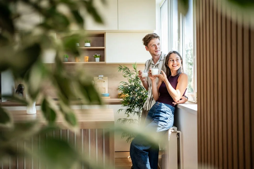 A man and woman enjoying coffee at a kitchen window, smiling and relaxed, with greenery in the foreground.
