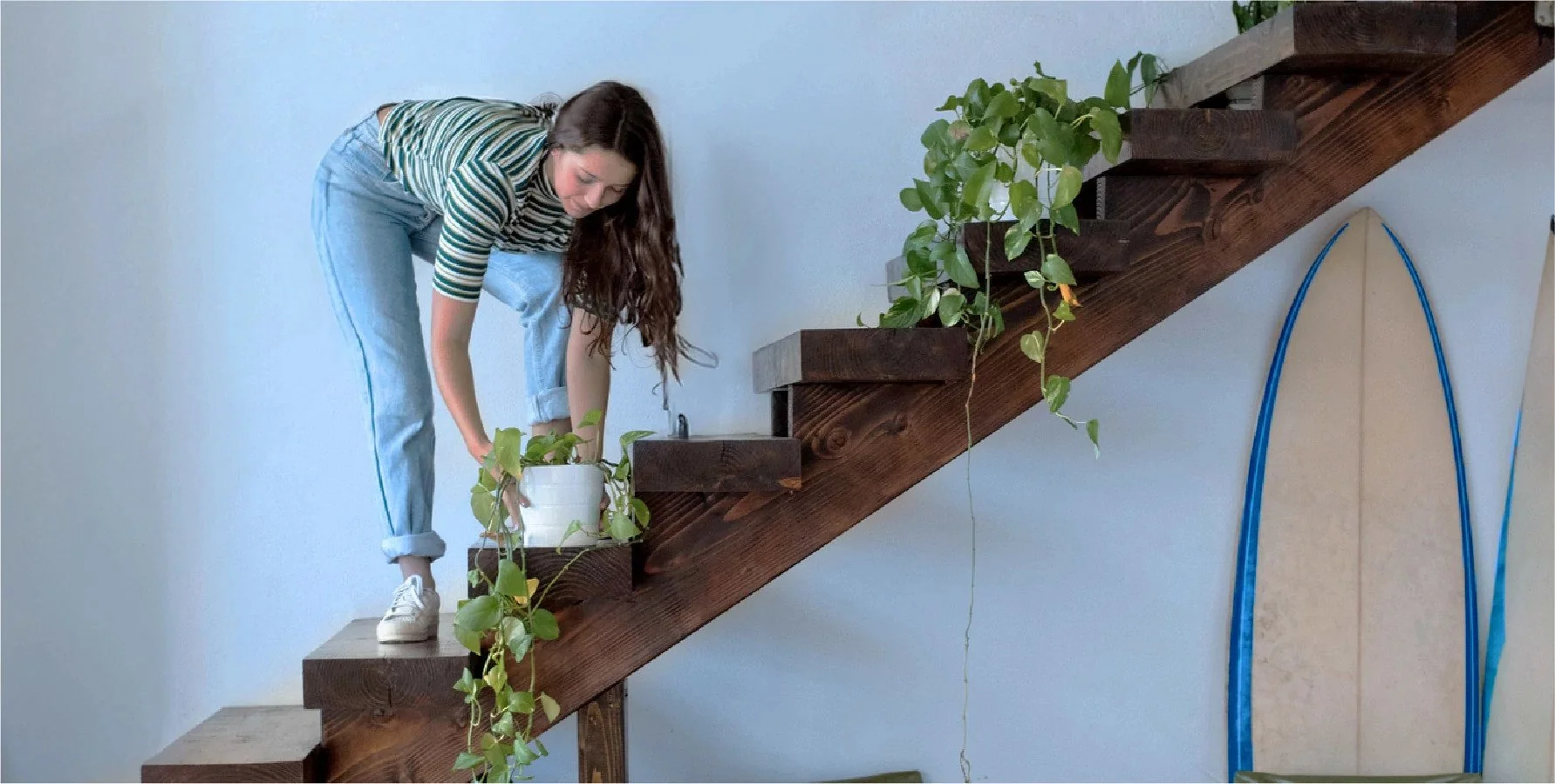 A woman in a striped shirt and jeans is on a wooden staircase, tending to a potted plant. The staircase has trailing vines, and surfboards are leaning against the wall nearby.
