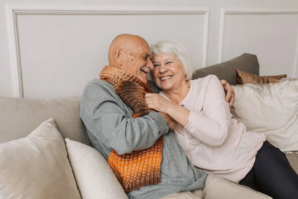 An elderly man and woman smiling and laughing together on a couch, embracing each other affectionately.