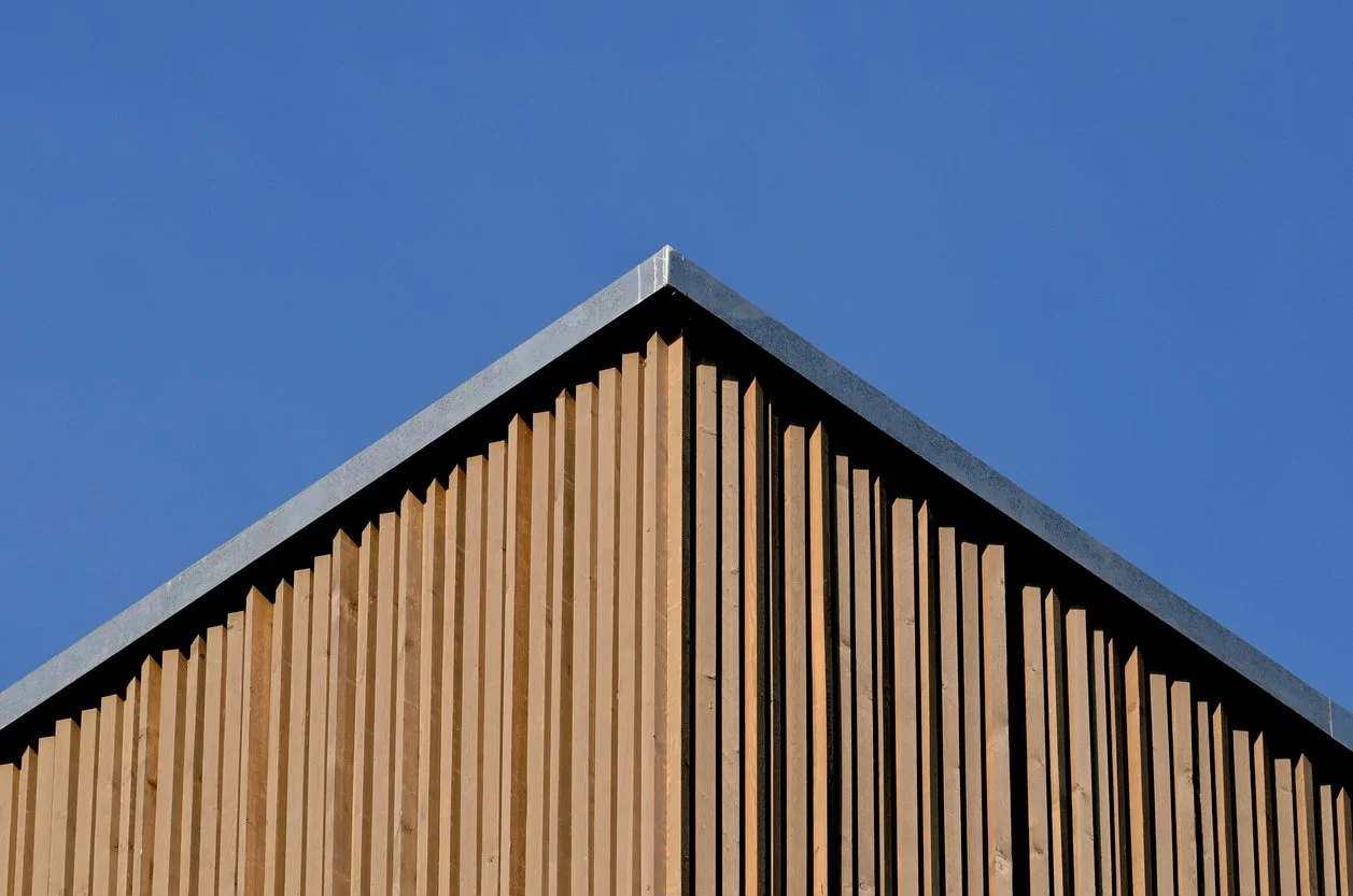 Close-up of the corner of a building with vertical wood slats and a metal roof, against a clear blue sky.