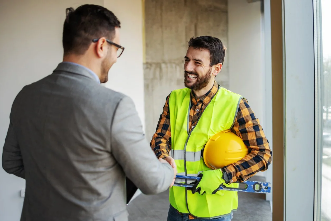Two men shaking hands indoors, one in a gray suit with glasses, the other in a yellow safety vest holding a yellow hard hat and a level, smiling and engaging in conversation near a large window.