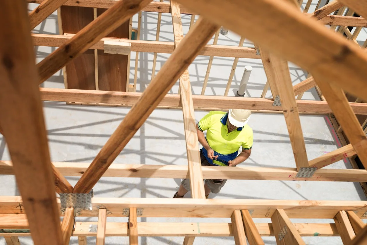 A construction worker wearing a white hard hat and neon yellow shirt is at a construction site, looking at a smartphone while working on a wooden framework.