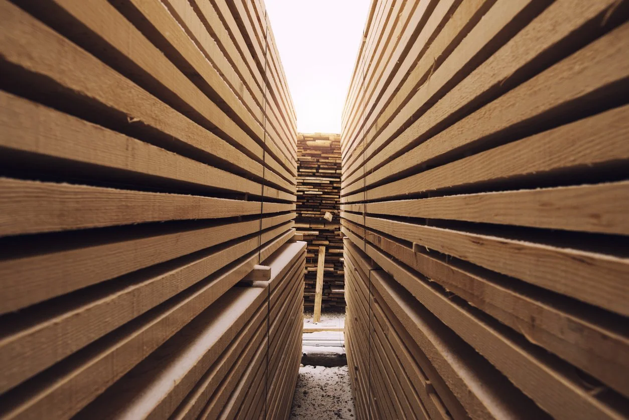 A narrow aisle between stacks of wooden planks outdoors, with the sky visible at the top.