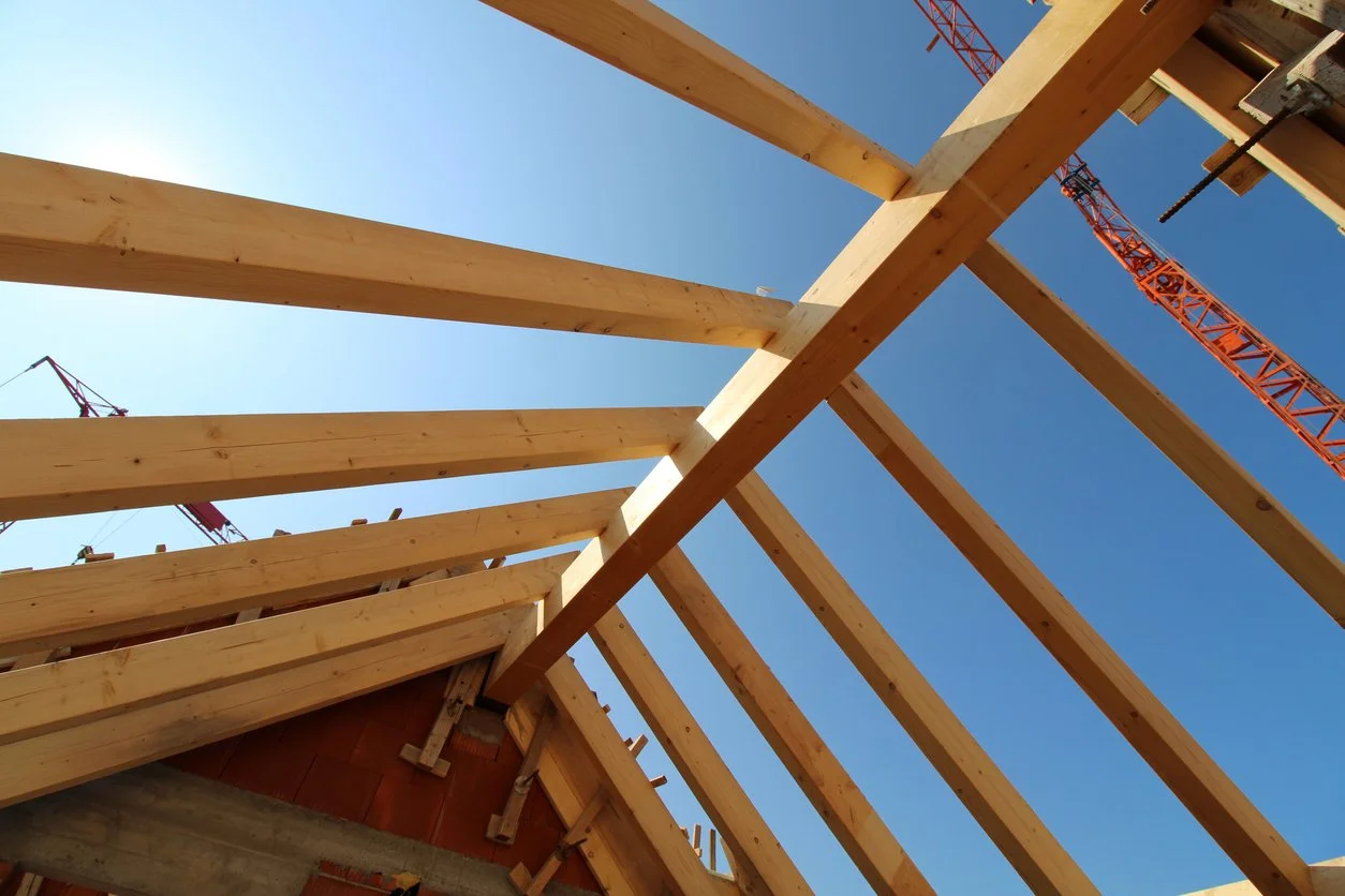 Construction site with wooden beams forming the roof framework, crane in the background, and a clear blue sky.