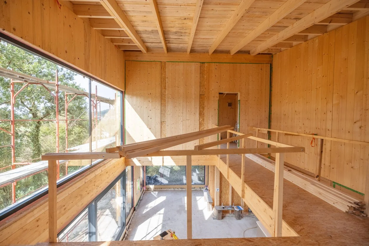 Interior view of a house under construction with wooden framing, walls, and ceiling. Large windows on the left side, construction tools and materials on the floor, and a partially built second story or loft area with wooden railings.