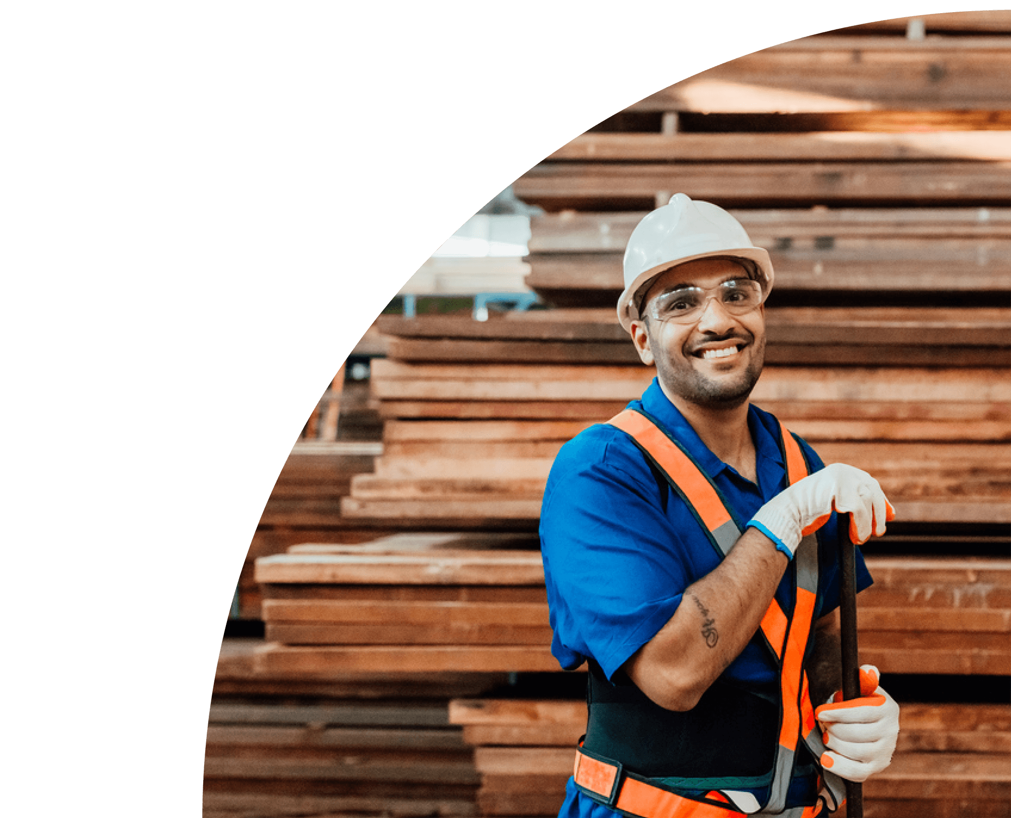 A construction worker wearing safety glasses, a white hard hat, and an orange safety vest smiling and holding a measuring tape in front of stacked wooden planks.