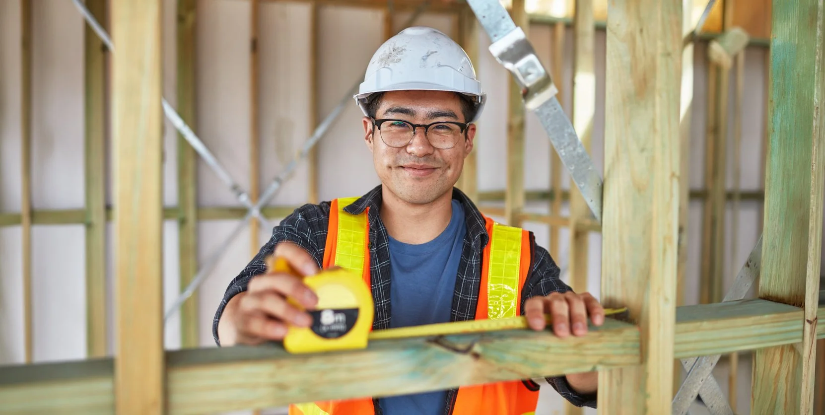Male construction worker wearing a white safety helmet, glasses, a blue shirt, and an orange safety vest, smiling while measuring wood framing with a yellow tape measure at a construction site.