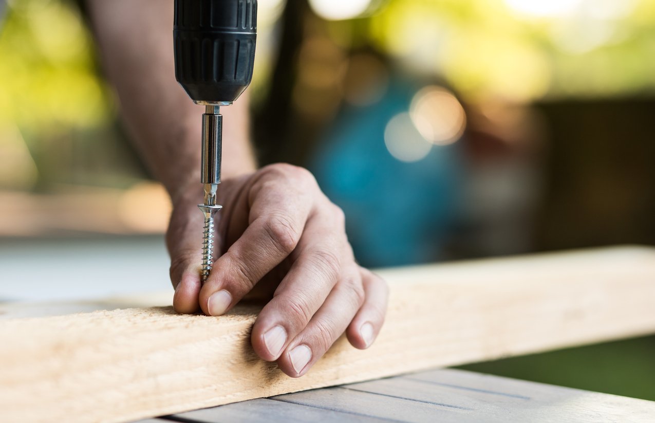 Person using a power drill to drive a screw into a piece of wood outdoors.
