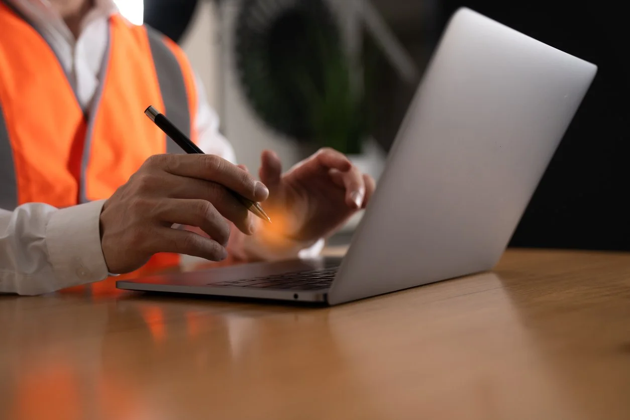 Person in safety vest writing on a laptop with a pen.