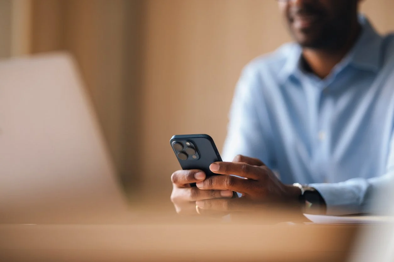 A man in a light blue shirt sitting at a desk, holding a smartphone in his hands, with a blurred laptop in the foreground.