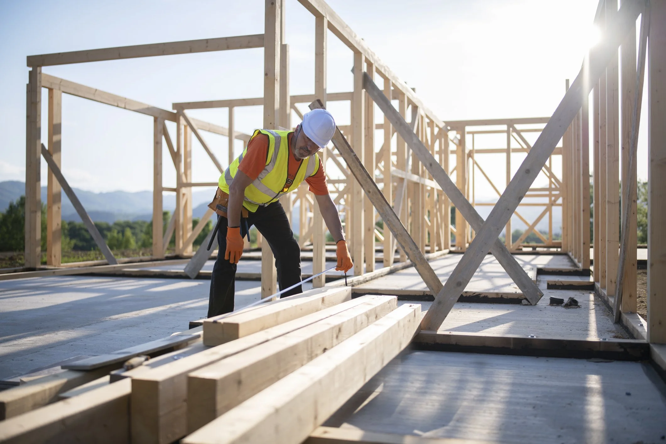 A construction worker wearing a white helmet and safety vest, working on a wooden framing structure at a building site during daytime.