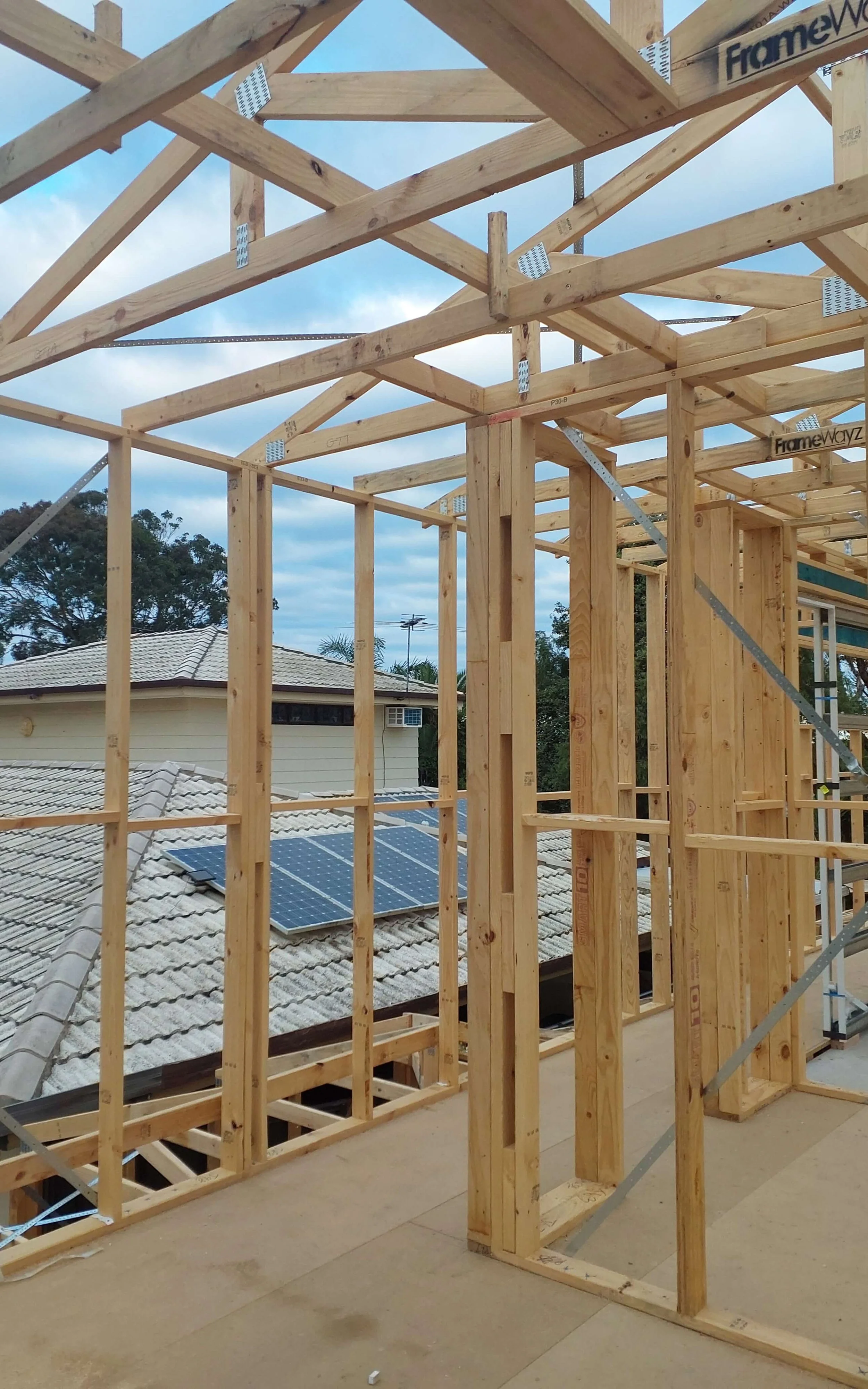 Construction site with wooden framing for a building, solar panels on neighboring rooftops, and a cloudy sky in the background.