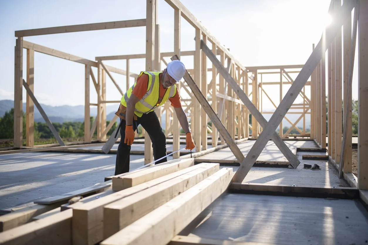 A construction worker wearing a white safety helmet, an orange shirt, a yellow safety vest, and orange gloves, inspecting wooden framing on a building site during daylight.