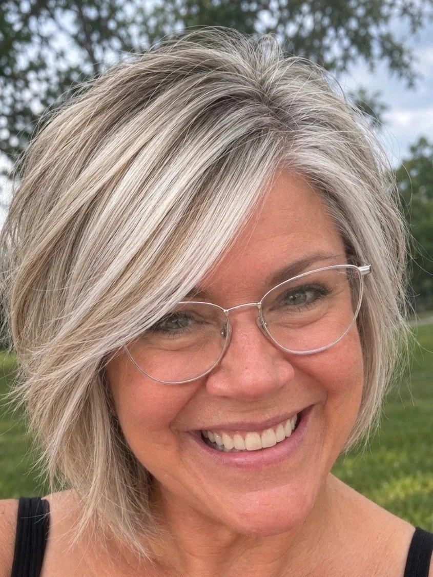 A woman with shoulder-length gray hair, wearing clear glasses and smiling outdoors with blurred trees in the background.