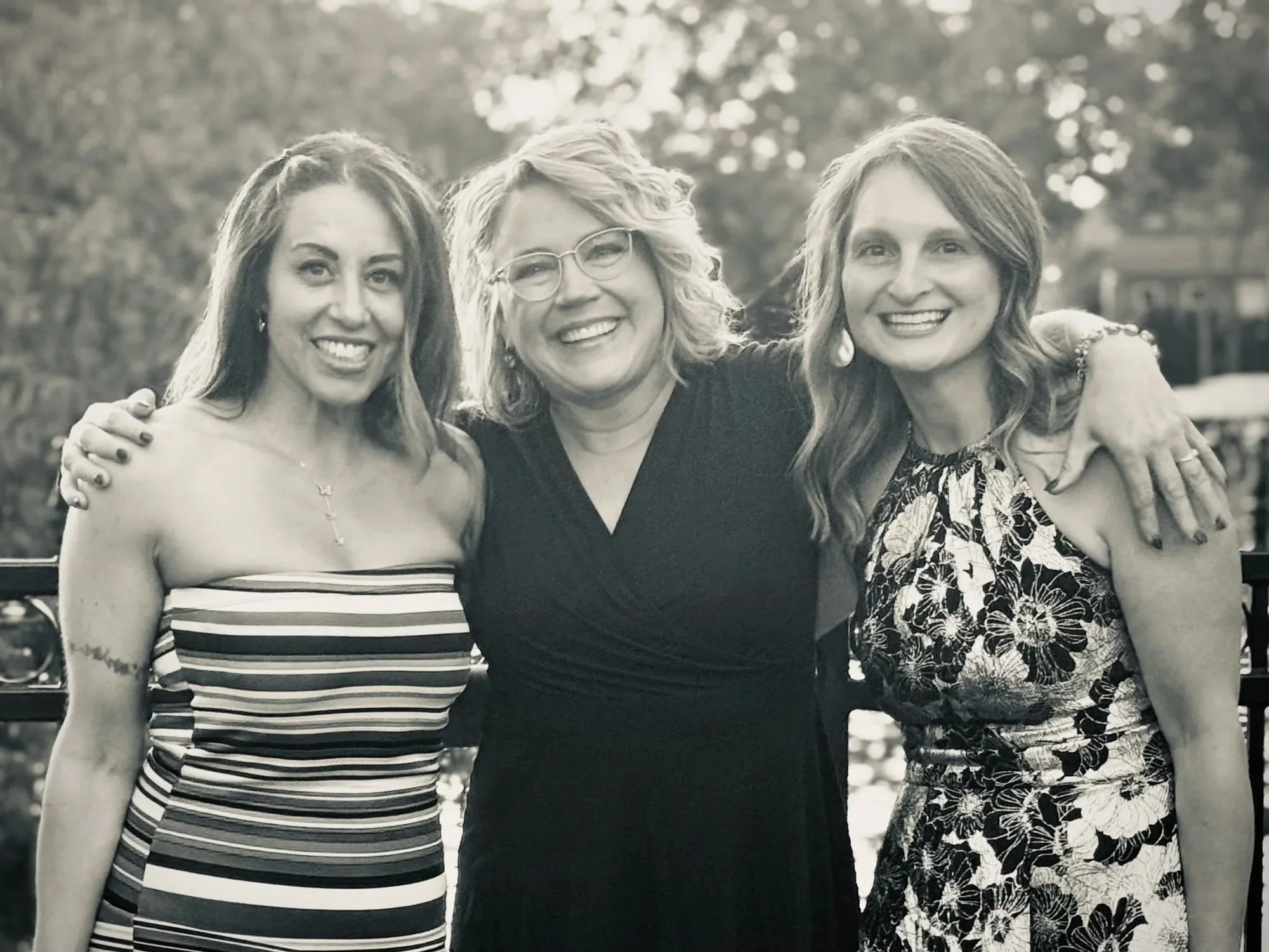 Three women smiling and posing outdoors, arms around each other, with blurred trees in the background.
