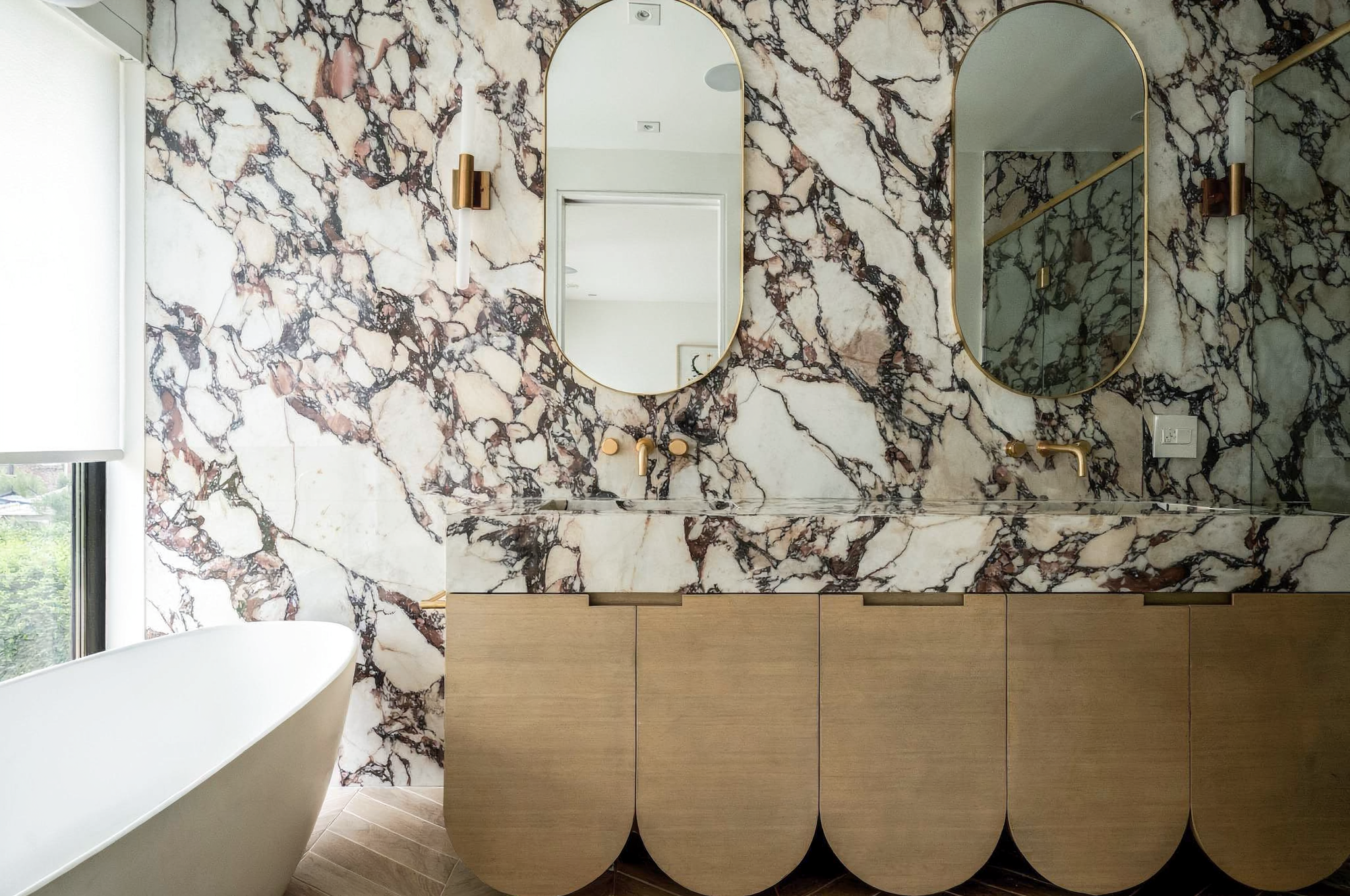 A bathroom with marble walls featuring black, white, and pink veining, two oval mirrors with gold trim, a white freestanding bathtub, and a wooden vanity with beige cabinets.