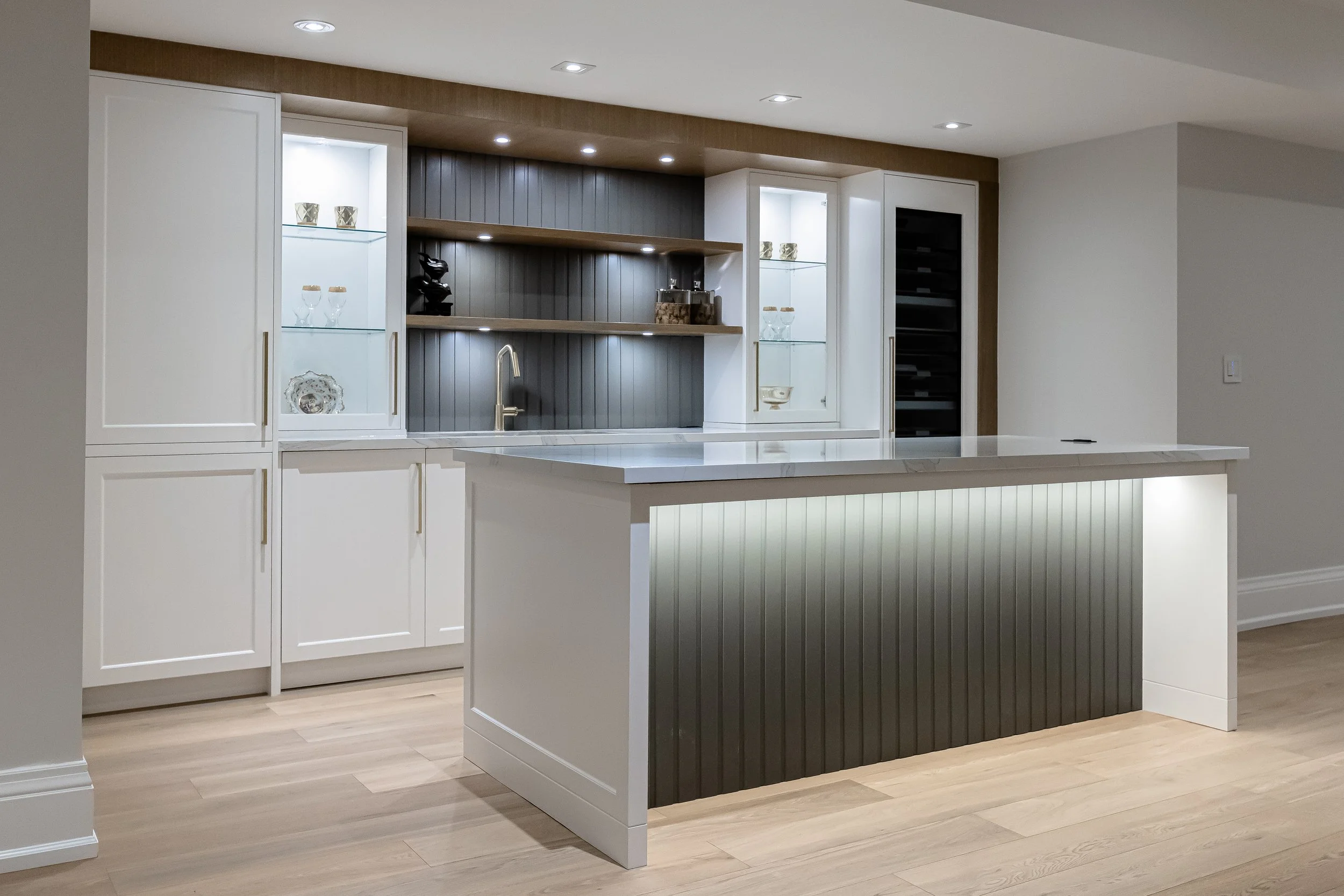 A modern kitchen with white cabinetry, an island with vertical paneling, and open shelves displaying decorative items and glassware. Recessed ceiling lights illuminate the space, and the floor is light wood.
