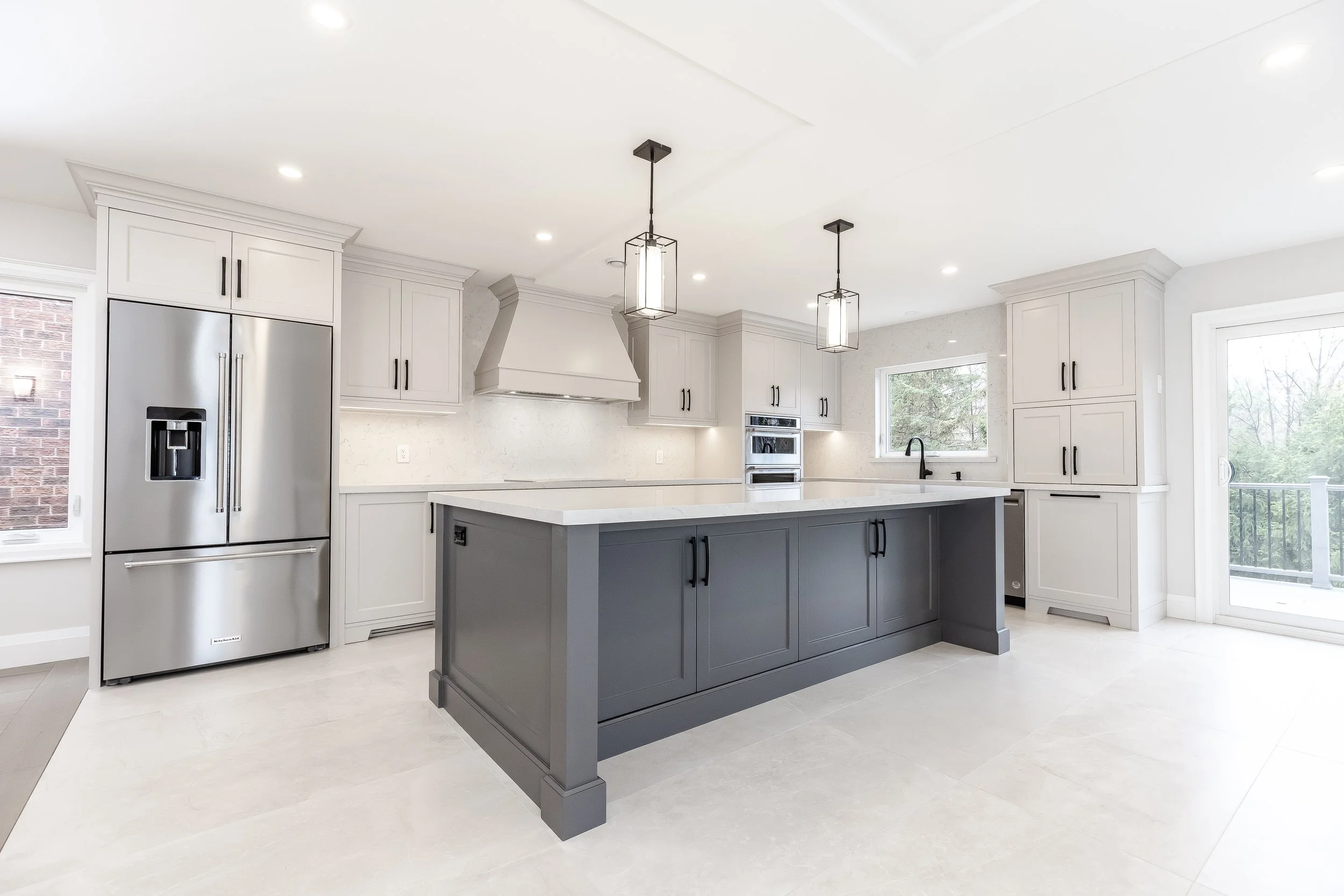 Modern kitchen with white cabinets, a gray island, stainless steel refrigerator, black fixtures, and large windows.