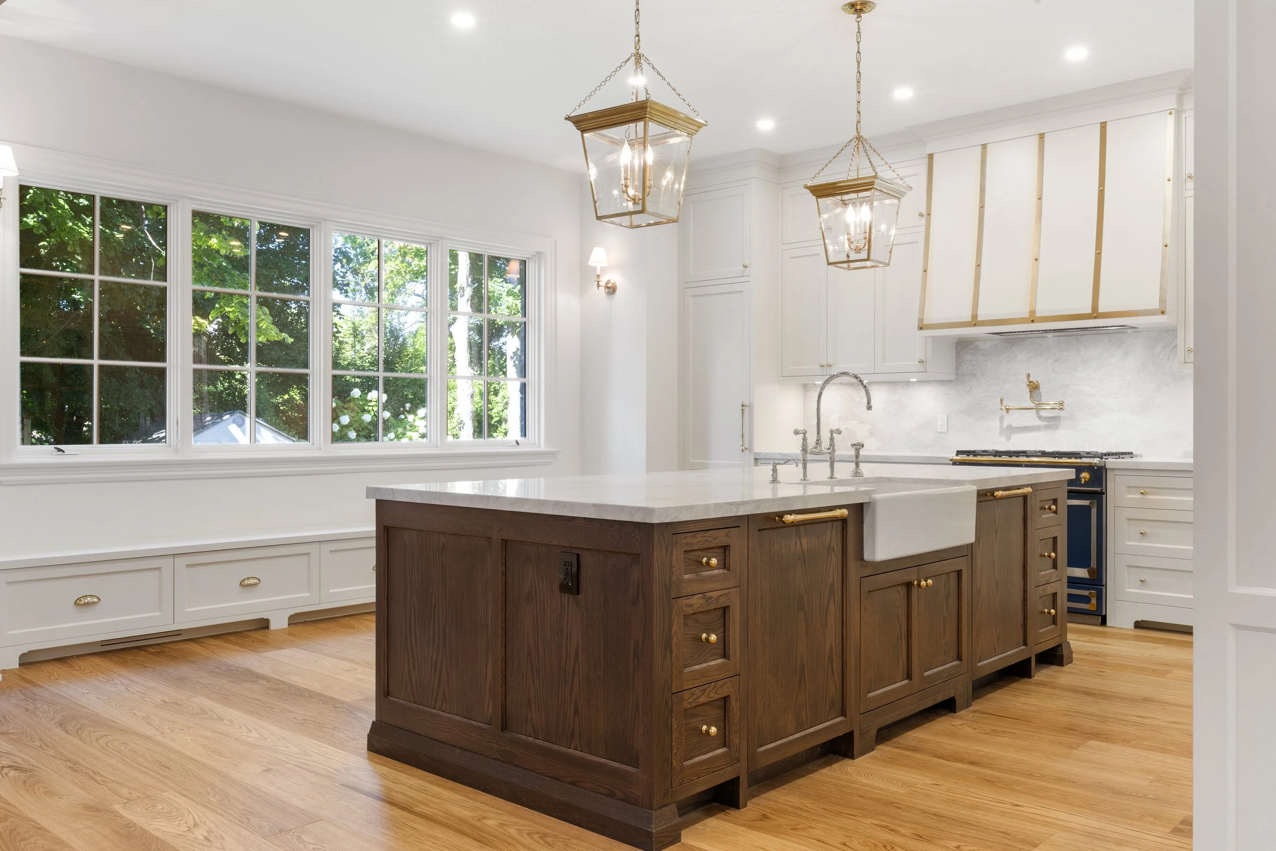 Modern kitchen with white cabinetry, a large window, a kitchen island with a marble top and dark wood finish, brass fixtures, and gold-accented hanging light fixtures.