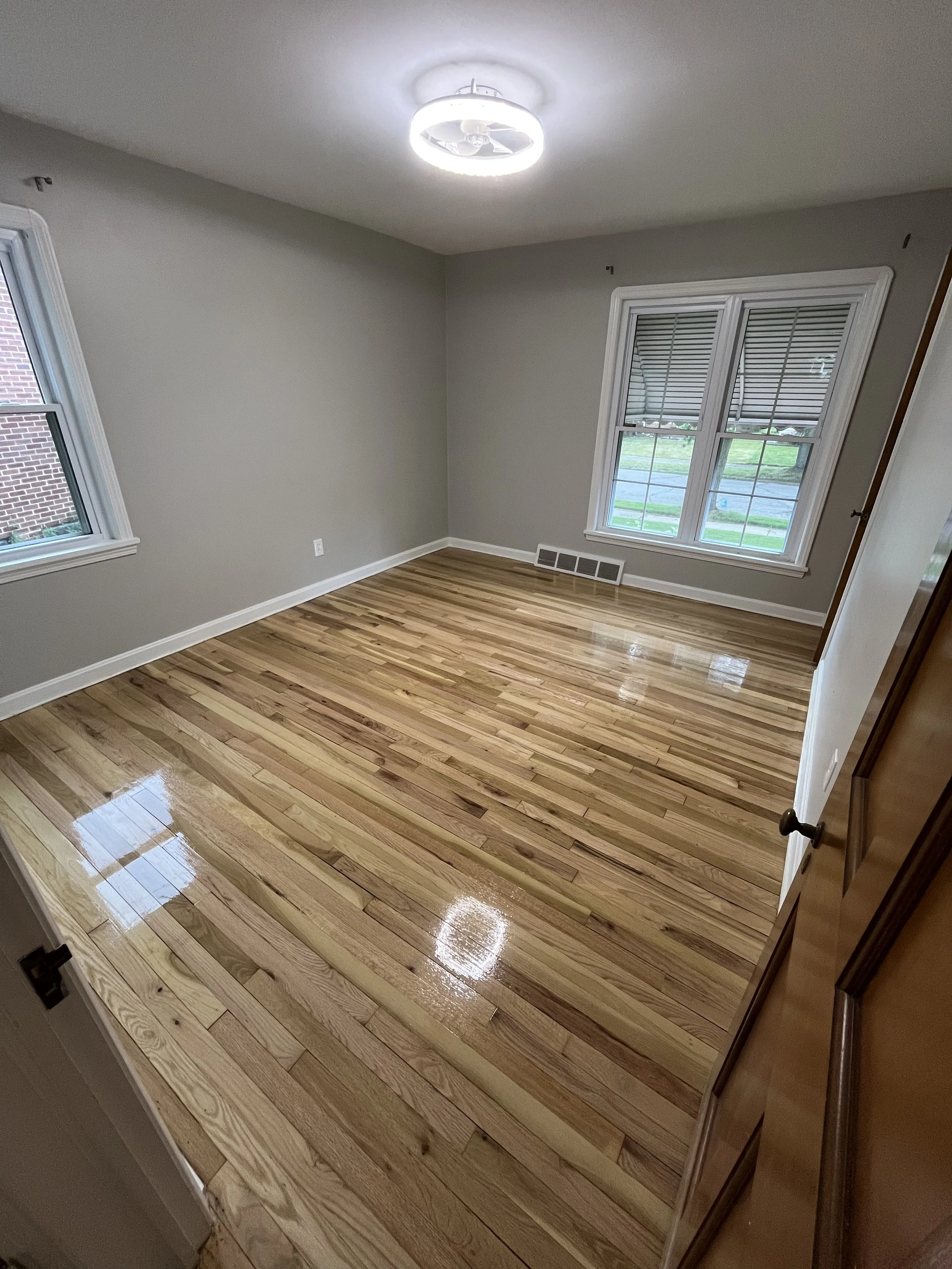 Empty room with hardwood floors, two windows with blinds, a ceiling light fixture, and a vent on the wall near the floor.
