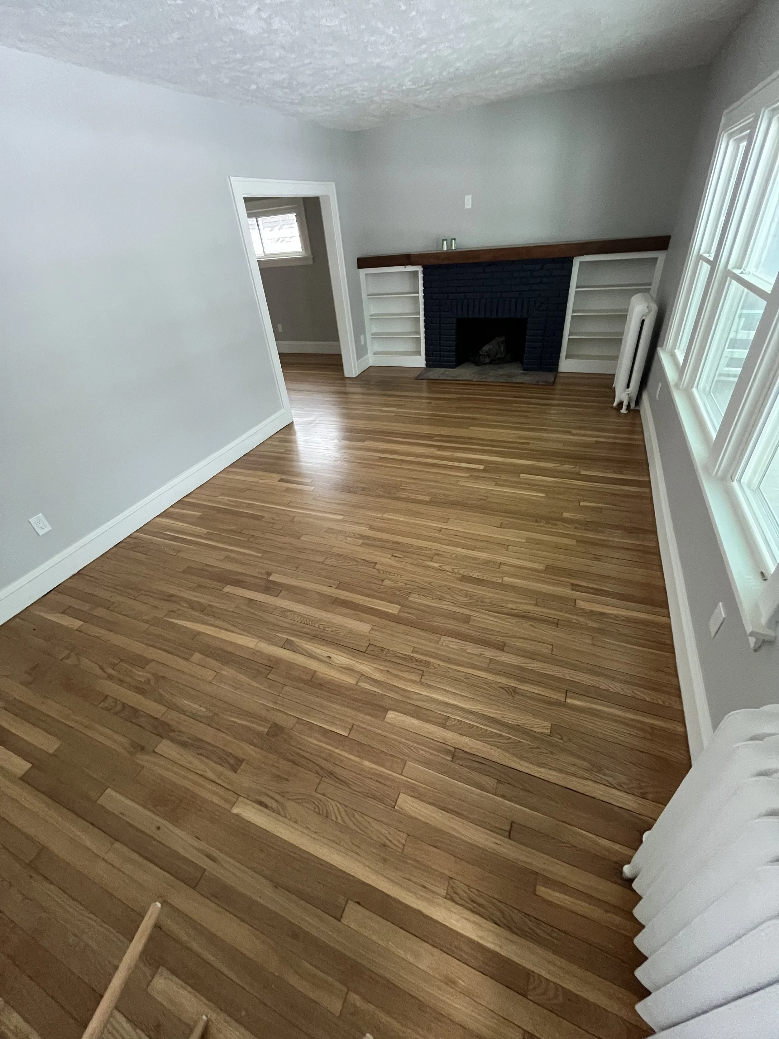 Empty living room with hardwood floors, a white fireplace with built-in shelves, a radiator, and large windows letting in natural light.