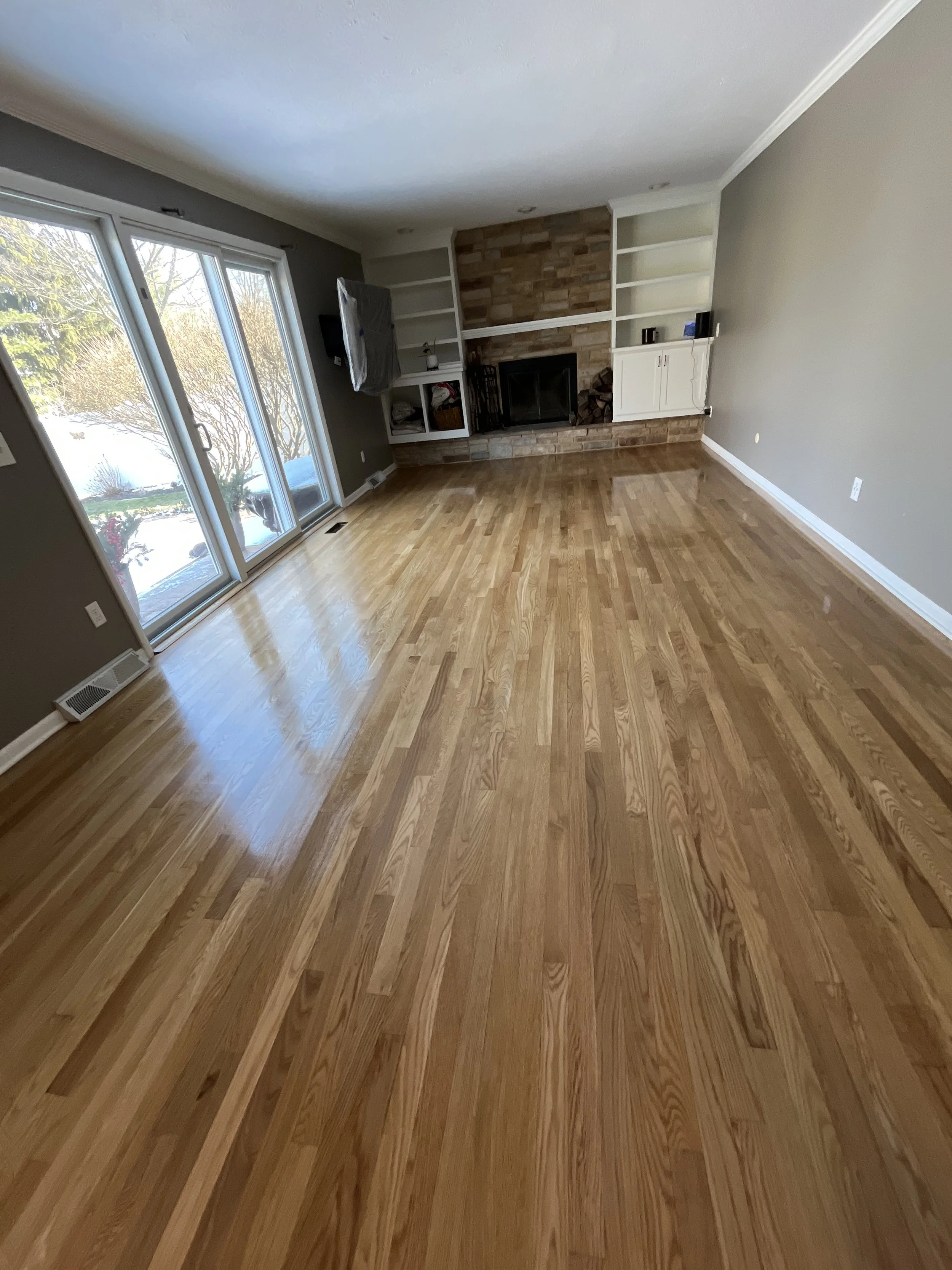 Empty living room with hardwood floors, a brick fireplace, built-in shelves, and a sliding glass door leading outside.