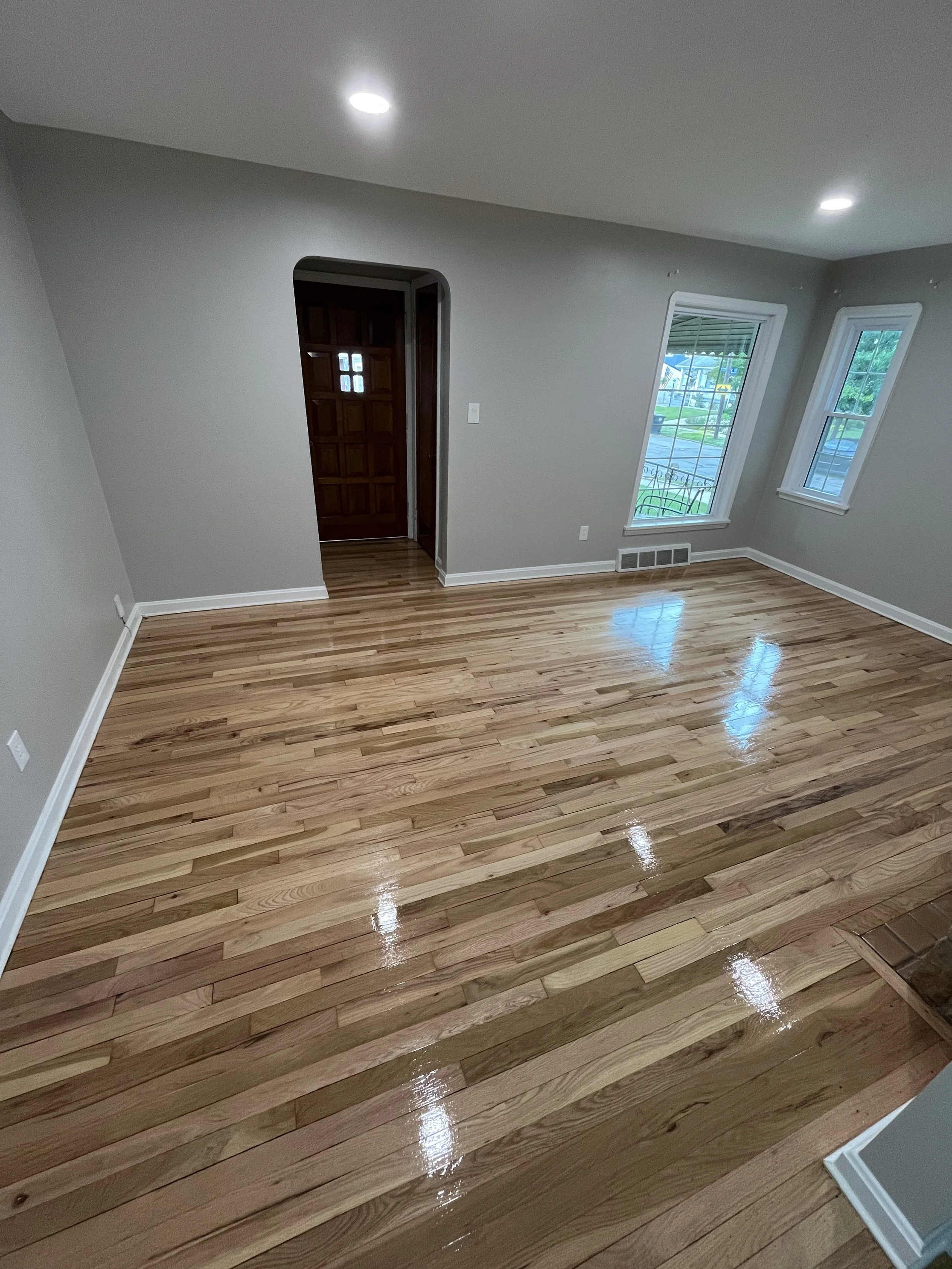 Empty living room with polished hardwood floors, two large windows letting in natural light, a wooden front door with small windows, and recessed lighting on gray walls.