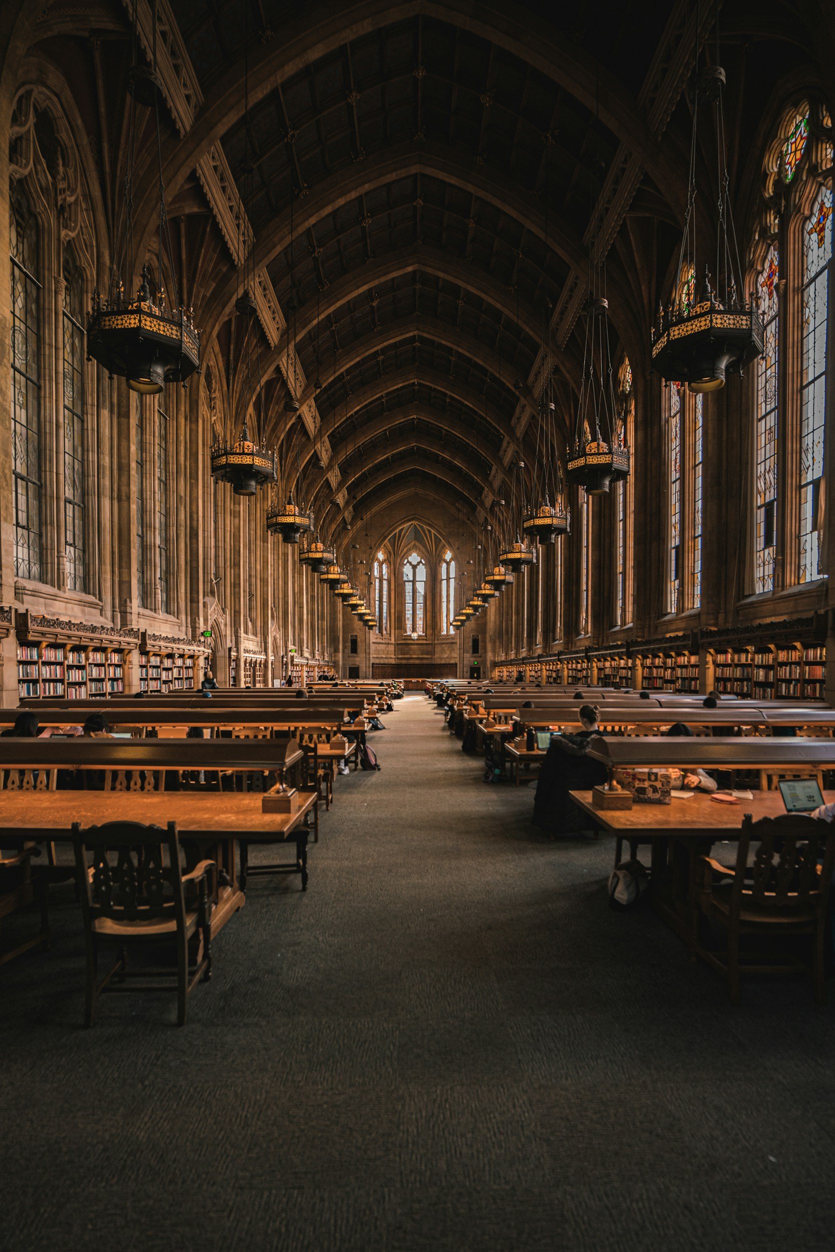 Interior of a historic library with high vaulted ceilings, stained glass windows, and rows of bookshelves filled with books. Wooden tables and benches are arranged for reading and studying, with some people seated and reading.